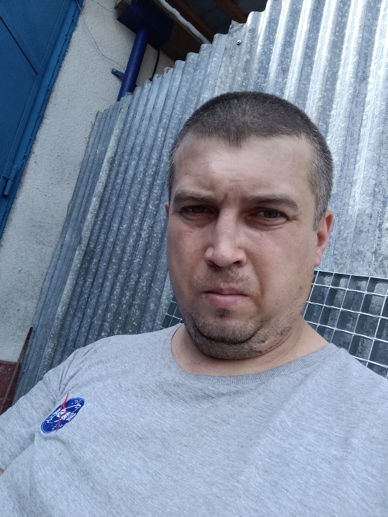 In Dalovy, Czechia, a man takes a selfie in front of a corrugated metal wall. He is wearing a grey t-shirt with a NASA logo on the left chest. The man has short, dark hair and a light beard. He is looking directly at the camera with a neutral expression. To the left of the man, a blue door is visible, and to his right, a metal grate is partially obscured. The overall impression is that of an outdoor setting, likely a yard or a utility area.