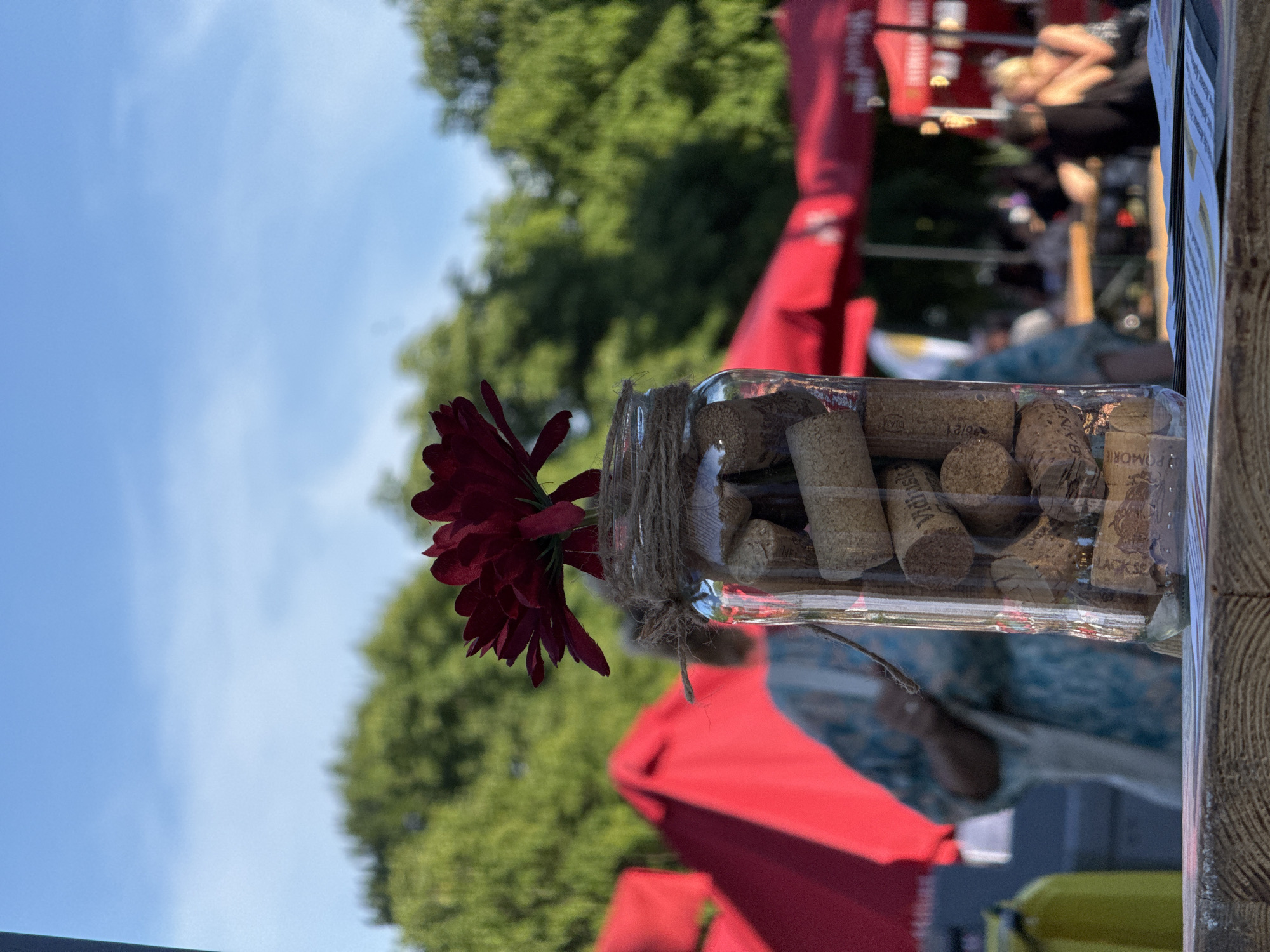 The image captures a close-up of a glass jar filled with wine corks, with a single deep red flower emerging from it. The jar is tied with twine, giving it a rustic decoration. Behind the jar, the scene is blurred, but suggests an outdoor event. Bright red canopies are visible, along with green trees and a clear blue sky, indicating a sunny day, likely in the late morning or afternoon. In the background, out-of-focus figures of people can be discerned, some appearing to be seated or interacting at what looks like a market or festival. The overall atmosphere seems relaxed and pleasant, fitting for an outdoor gathering. Some text is visible on the wine corks, including "VICTORIA" and "LAGI 32," suggesting they are from various wine brands. The wooden texture on the right edge of the frame hints at a table or display surface. The setting is likely an outdoor market or festival in Burgas, Bulgaria, given the provided location context.