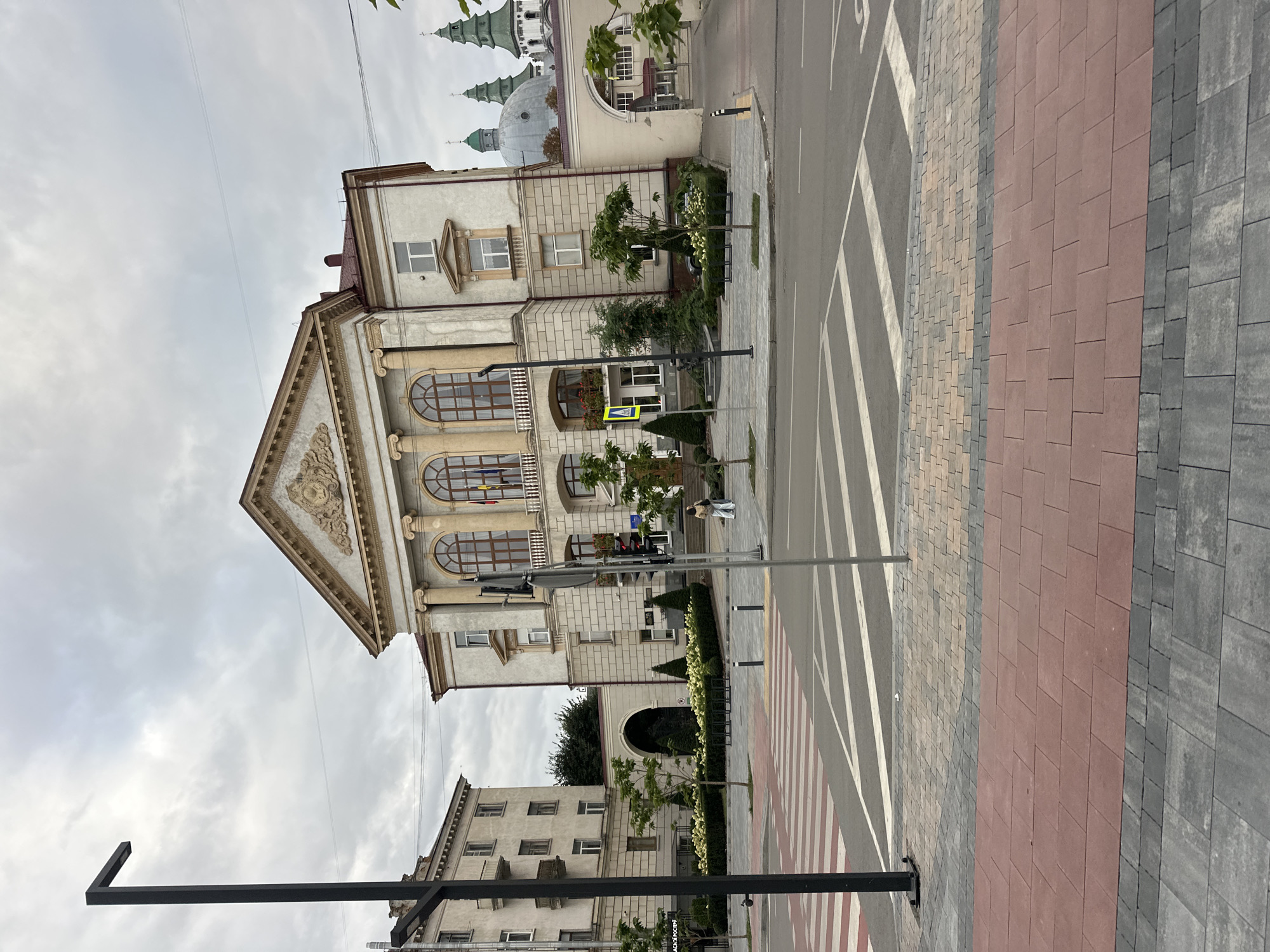 The image depicts a frontal view of the Ternopil Regional State Administration building in Ternopil, Ukraine. The building is a large, light-colored edifice with classical architectural elements, including columns, arched windows, and ornate friezes. In front of the building, there is a well-maintained plaza with trees, bushes, and benches. To the left of the main building, another multi-story apartment building is visible. The foreground shows a pedestrian crossing with red and white stripes, and a street with markings. The sky is overcast. A single person is standing in the middle ground, facing away from the camera, near the entrance of the administration building. There are streetlights and traffic signals present. The overall scene conveys an urban setting with civic architecture.