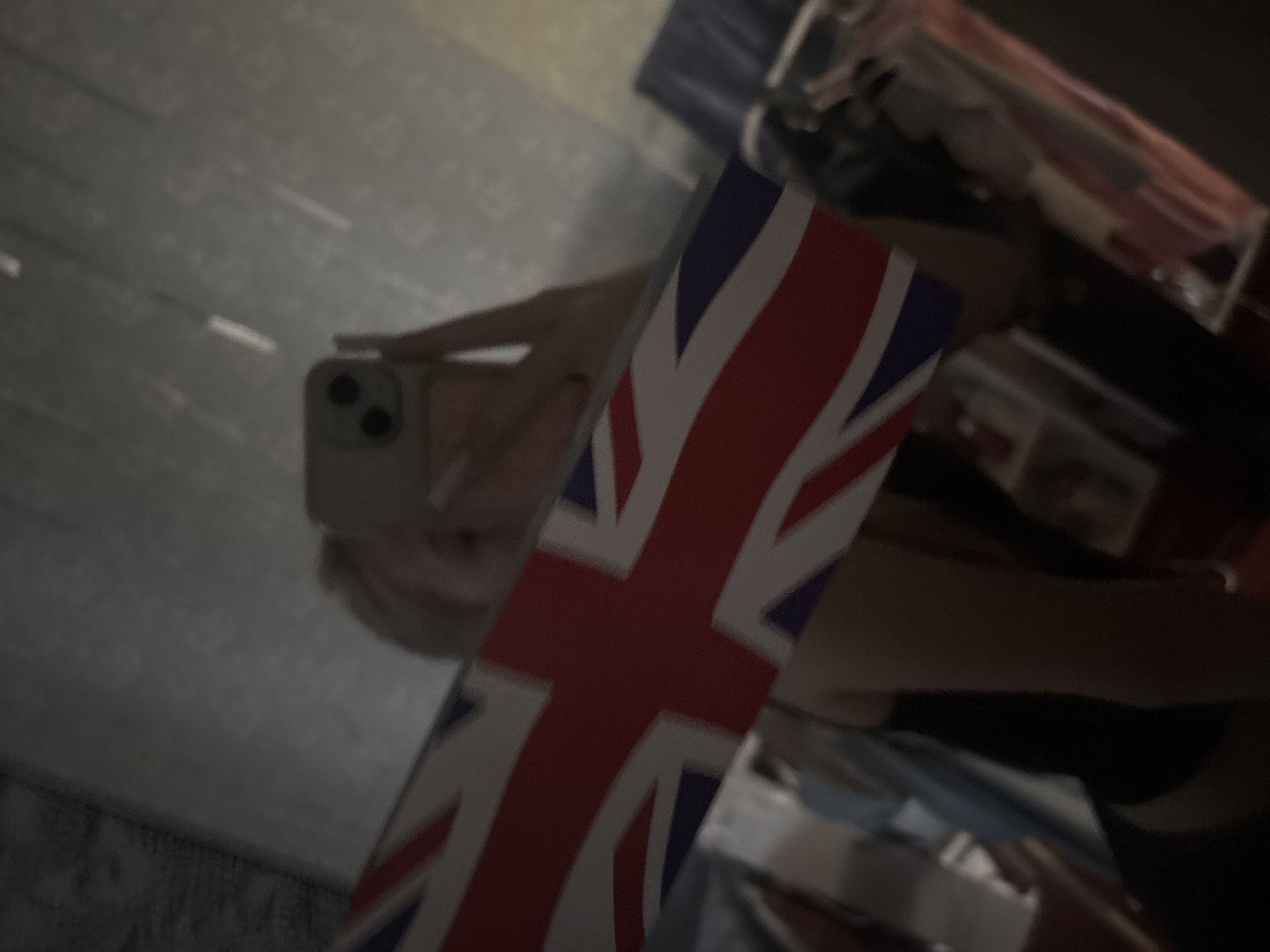 A young woman, her face obscured by a reflection in a mirror, holds up a small Union Jack flag. She is wearing a black top and her blonde hair is visible. The reflection shows her holding a smartphone and a portion of her body. The background of the mirror is dark and textured, with some light streaks. The scene appears to be indoors, possibly in a bedroom or dressing area. The image is blurry and lacks clear details of the environment or any specific location within Velykyi Sukhodil, Ukraine.