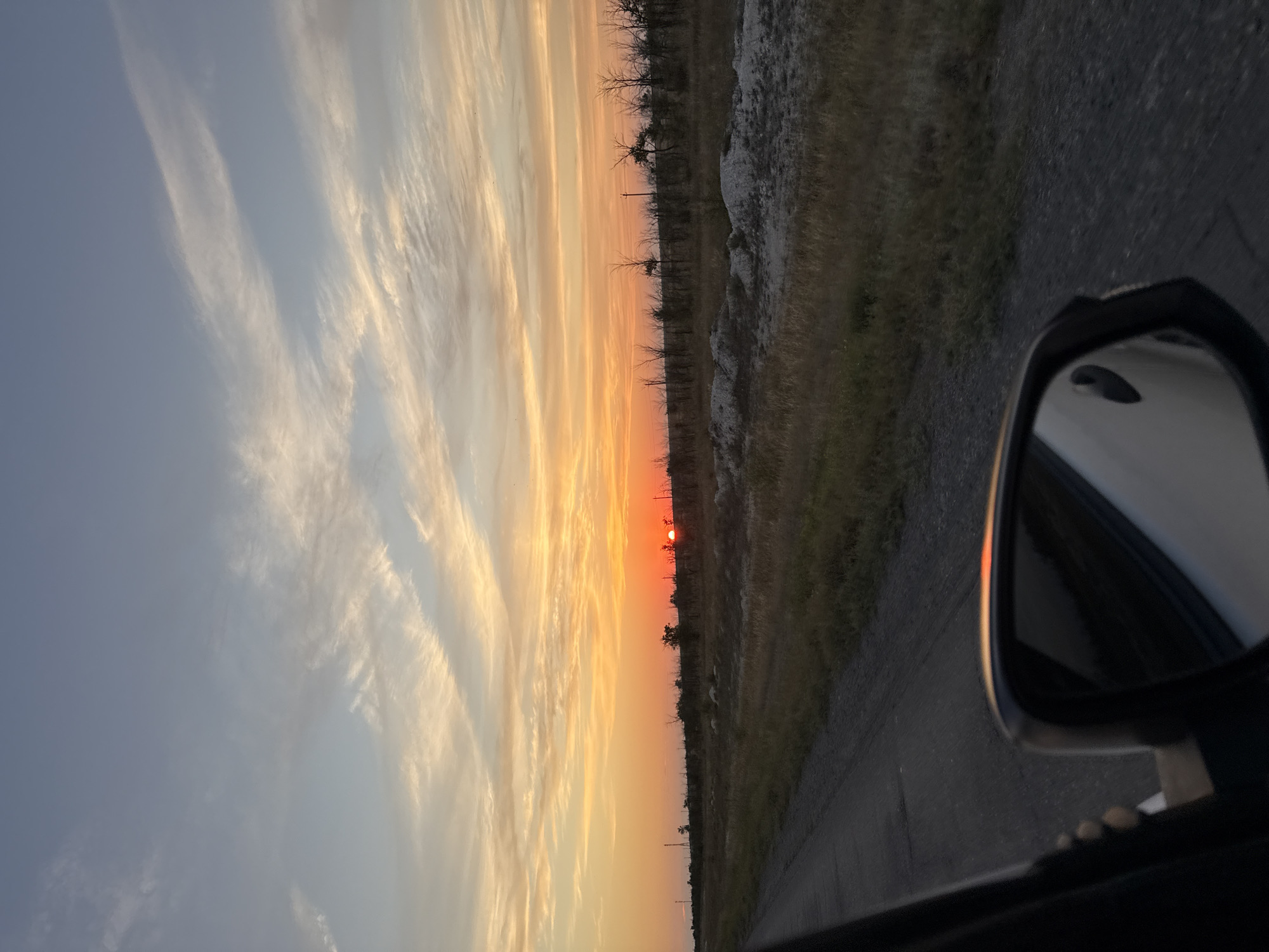The image captures a sunset over a rural landscape, viewed from inside a vehicle. The sky is painted with vibrant hues of orange and yellow, transitioning to soft blues and whites, with wispy clouds streaking across the horizon. The sun, a bright orange orb, is partially visible, casting a warm glow over the scene. In the foreground, a side-view mirror of the car reflects the sky and the interior of the vehicle. A road, likely paved with asphalt, runs alongside a dry, grassy field dotted with sparse, leafless trees. The landscape appears flat and open, typical of the rural areas of Пугачёв ауылдық округі, Kazakhstan. The overall impression is one of a peaceful, twilight journey through the Kazakh countryside.