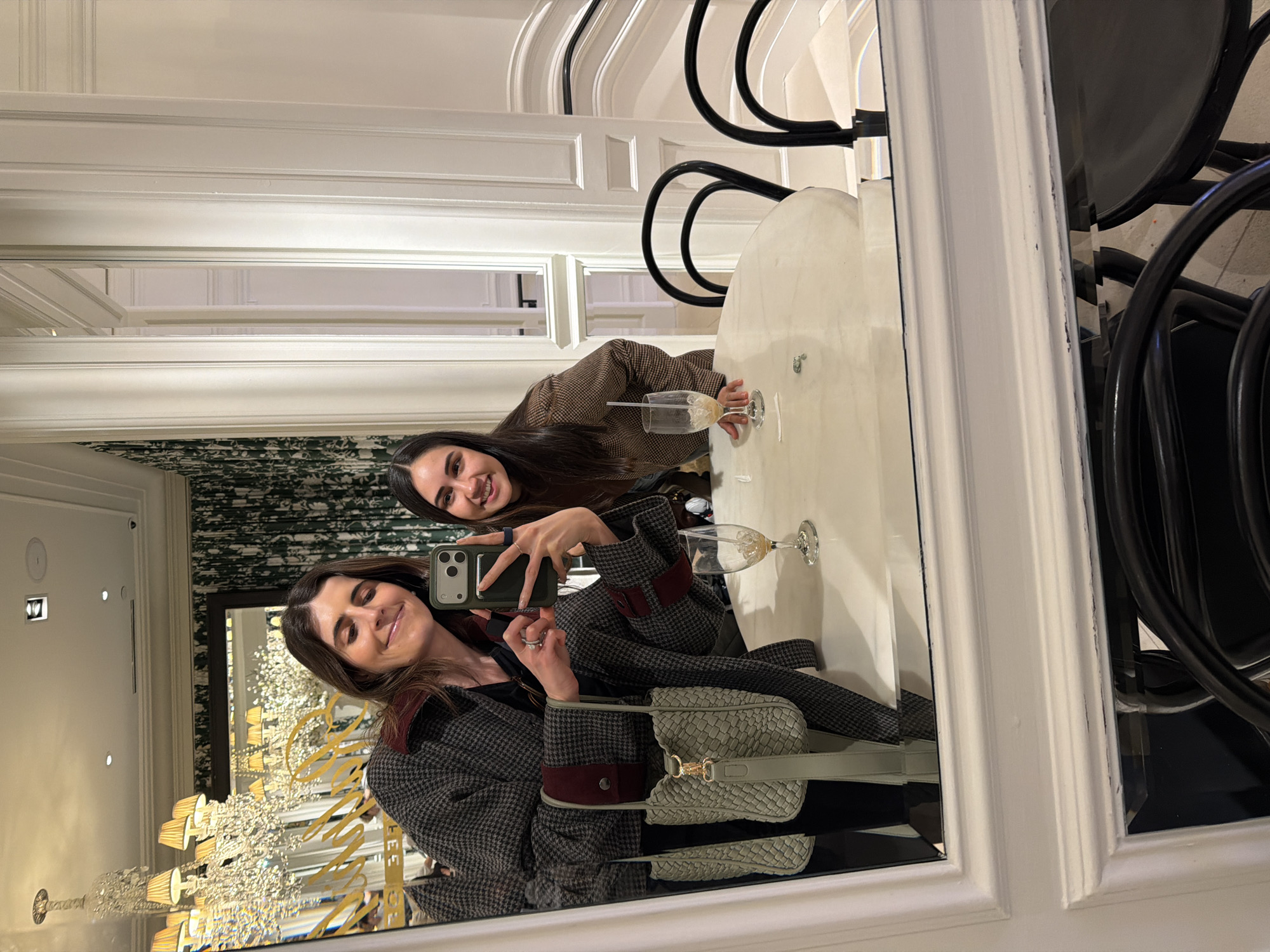 Two women are captured in a selfie reflected in a large mirror within an opulent interior, likely a cafe or restaurant inside Bergdorf Goodman, New York City, United States of America.
The woman on the left, holding a dark green smartphone with a dark green case to take the photograph, has brown hair and is smiling. She wears a grey and black tweed-patterned coat with dark red lapels and cuff details, and a black garment underneath. A light green woven bucket bag with a gold clasp hangs from her shoulder. She wears rings on her left index and ring fingers. The woman on the right, partially visible, also has dark brown hair and is smiling. She wears a brown patterned jacket.
In the foreground, two black Thonet-style chairs are pulled up to a white oval table. On the table are two empty wine glasses; one contains residual light brown liquid and a white straw.
The background, reflected in the mirror, features elaborate decor. A large, multi-tiered crystal chandelier with numerous lampshades illuminates the scene. Dark green wallpaper with a lighter patterned motif covers the upper walls. White classical wall paneling and moldings frame the mirrors and walls. Gold script lettering, clearly reflecting "BERGDORF" and partially "THE PALM", is visible on a wall or mirror. A white staircase with black handrails ascends in the background on the right.