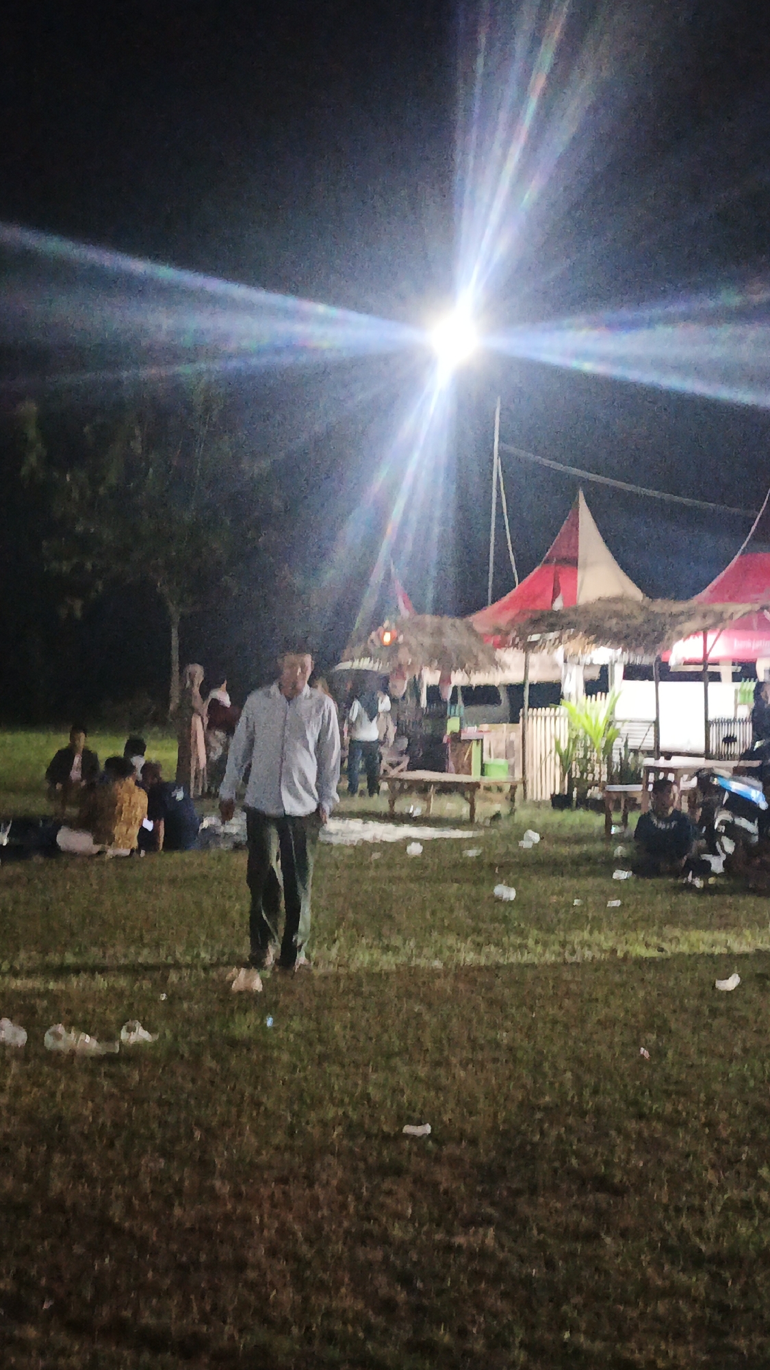 The image depicts an outdoor gathering at night in Ketemasdungus, Indonesia. A bright light source in the sky creates a starburst effect, illuminating a grassy area where several people are present. In the foreground, a man in a light-colored shirt and dark pants walks across the lawn. In the background, two tents with red and white canopies are set up, appearing to be stalls or serving areas, with thatched roofs. People are scattered around the tents, some seated on the ground and others standing. There are scattered pieces of litter on the grass. Trees are visible in the darker areas of the background.