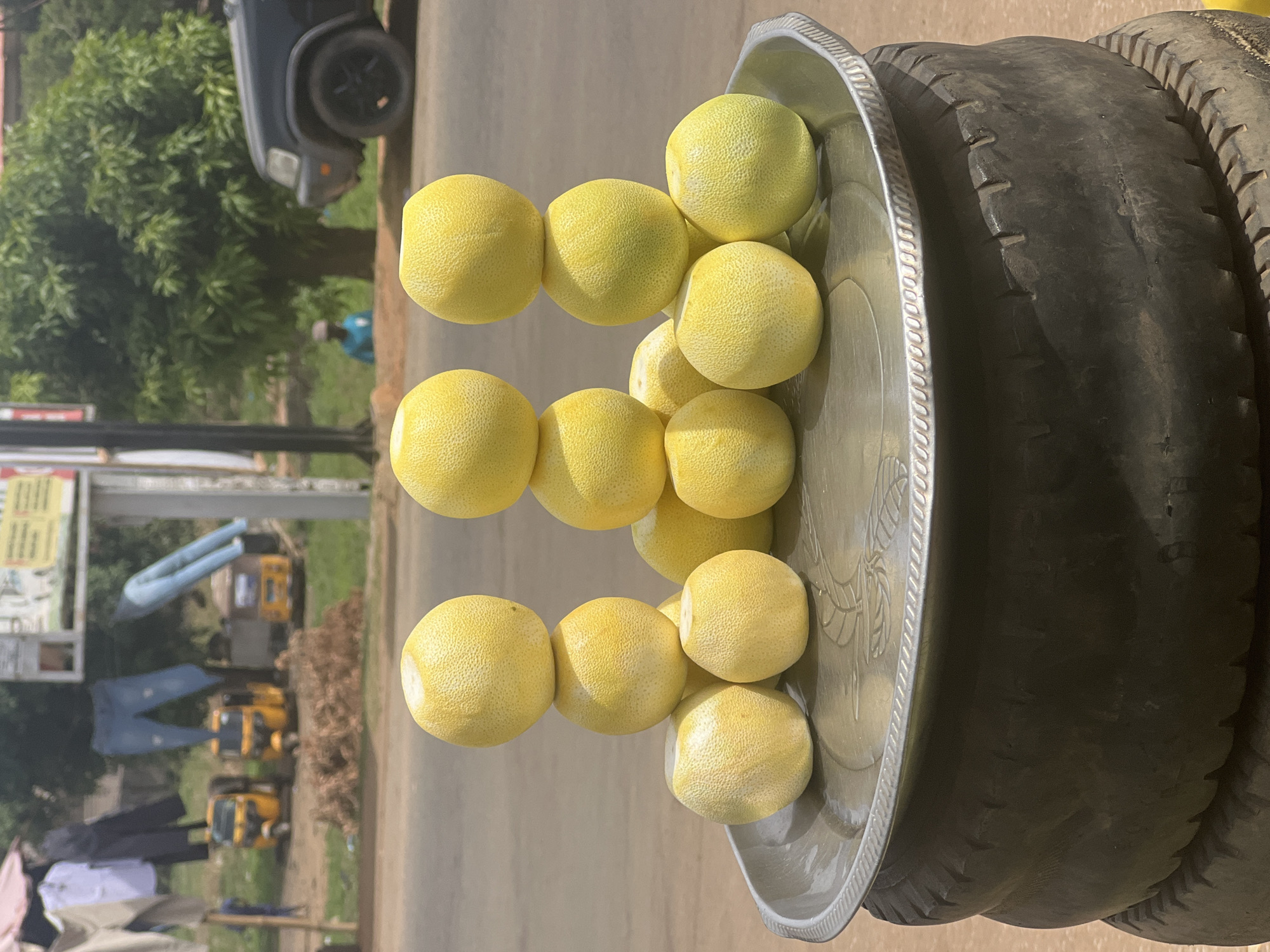 The image, captured in Galadimawa, Nigeria, presents a vibrant outdoor scene focused on a street vendor's display of fresh produce. The photograph is rotated 90 degrees clockwise, showing a brightly lit roadside setting.
The main subject is a large, round, silver-colored metal tray laden with numerous light yellow, spherical fruits, likely lemons or small pomelos. These fruits have a textured, bumpy skin and are neatly stacked in three columns, with additional fruits visible within the tray. The tray itself features an ornate, embossed rim and a faint pattern resembling wheat stalks etched onto its surface. This display tray is resting on a sturdy base comprised of at least two stacked, dark, worn vehicle tires.
In the background, to what would be the left side when oriented correctly, a paved road is visible. A dark-colored vehicle, possibly an SUV or pickup truck, is partially seen on the road. Behind it, lush green trees and foliage line the street. Further down the road, several yellow three-wheeled auto-rickshaws (tuk-tuks) are parked or in transit. Clothes, including a prominent pair of blue jeans, are draped over rails or lines, suggesting either items for sale or laundry drying. A blurred sign or banner with indecipherable text is also visible on the far left. A single person, wearing a blue shirt, is faintly visible among the trees and vehicles in the distance, though their activity is unclear.
The scene depicts a static arrangement of goods, indicating a vendor's stall where fruits are offered for sale. No direct activity of buying or selling is captured. The bright, direct sunlight suggests it is daytime, likely midday or early afternoon, evidenced by the distinct shadows. The overall environment is consistent with a bustling, informal market or roadside setup common in regions like Galadimawa, Nigeria. There is no clearly readable text on any visible signs or objects within the frame.