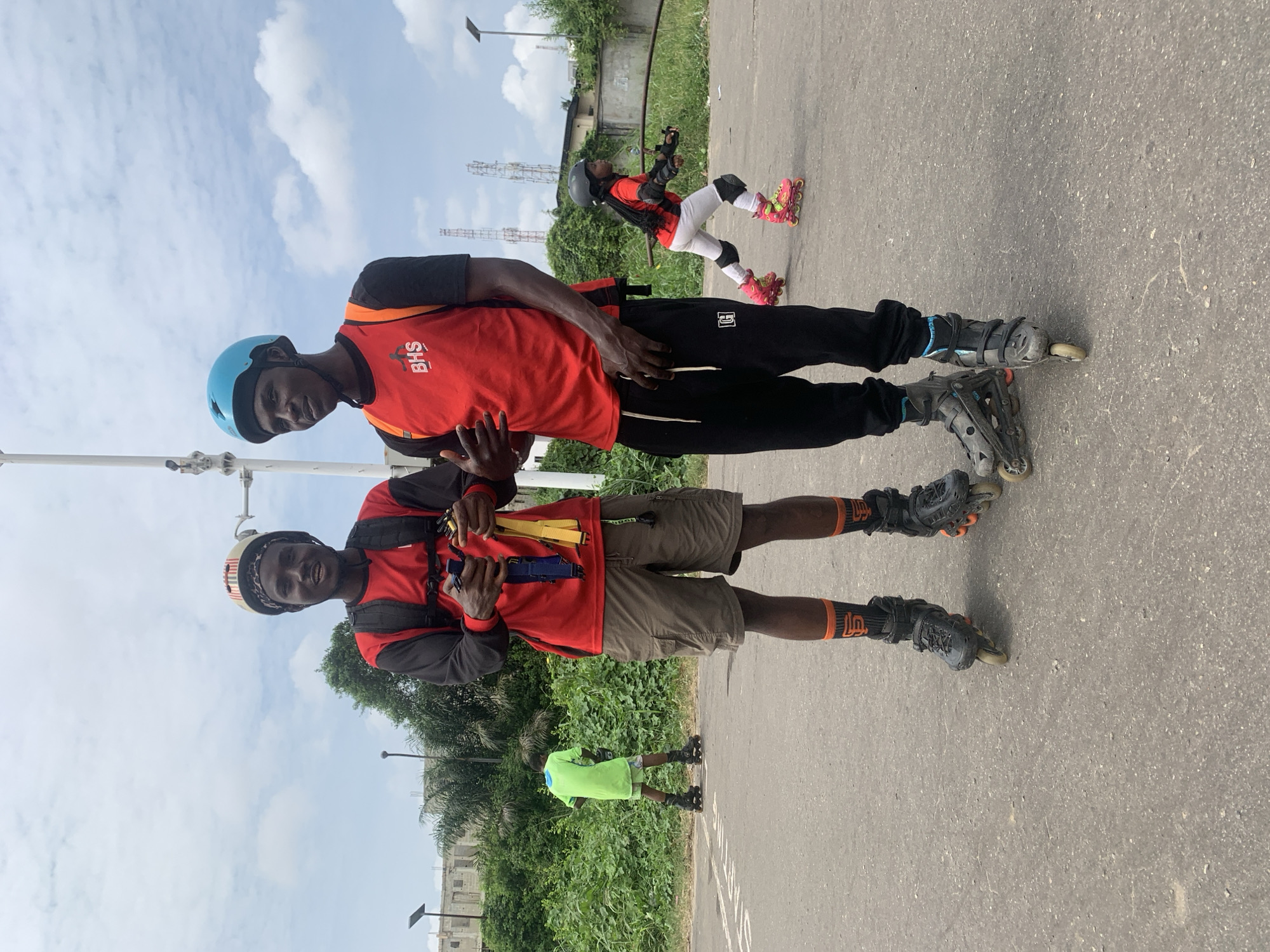 In Alagbede, Nigeria, two young men are posing on an asphalt road, both wearing rollerblades. The man on the left has a striped helmet and a red long-sleeved shirt with a black vest. He is holding what appear to be rollerblade adjustment tools in his hands. He is wearing khaki shorts and orange and black patterned socks. The man on the right is wearing a blue helmet and a red vest over a black t-shirt. He is wearing black pants and dark rollerblades. In the background, another young person in a bright green shirt and shorts is also rollerblading, and a young girl in protective gear and pink rollerblades is further in the distance. Lush green vegetation lines the road, and various structures, including buildings and communication towers, are visible in the background under a cloudy sky.
