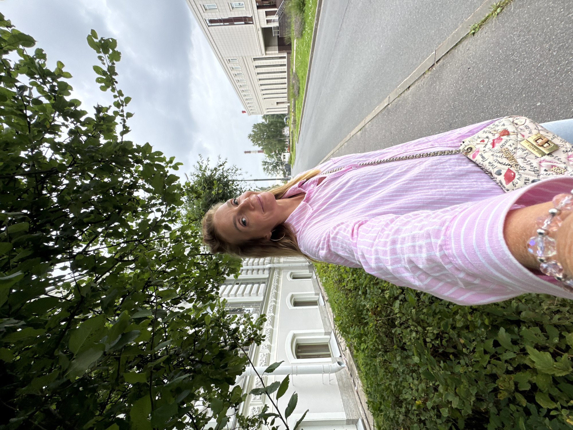 A woman with blonde hair, wearing a pink and white striped shirt and a bracelet on her left wrist, is taking a selfie outdoors. She is standing on a sidewalk next to a street. To her left is a building with white walls and windows, and a row of green bushes. To her right is the street, with a modern building in the distance. The sky is overcast. The location is Ladozhskoe Ozero, Russia.