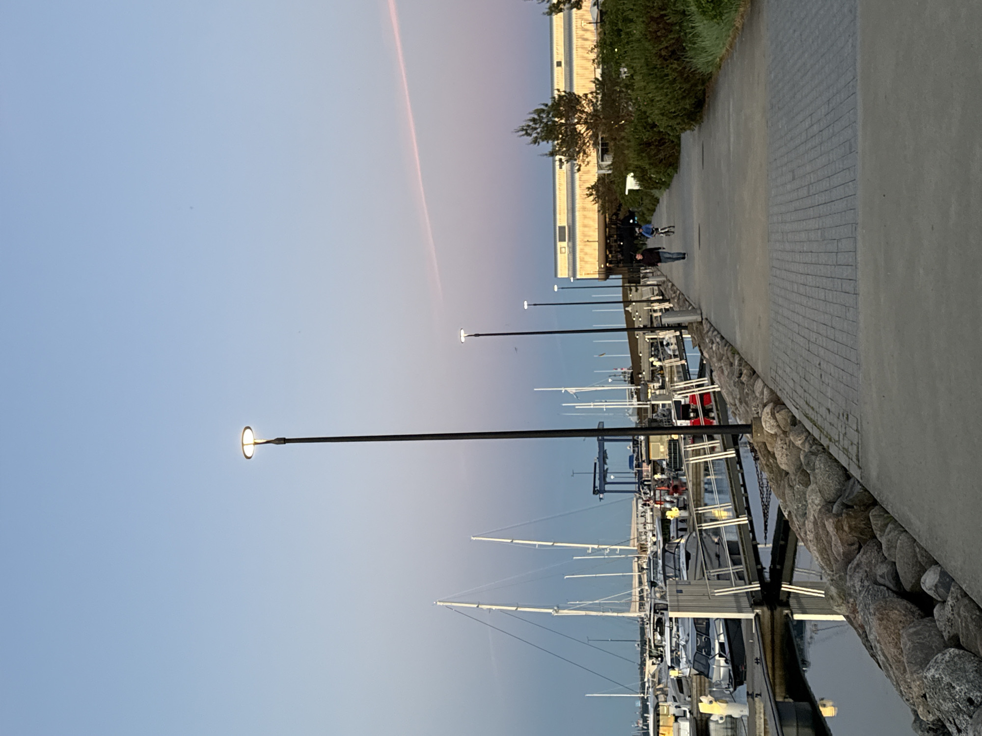 The image depicts a harbor scene in Tallinn, Estonia, at dusk. A paved walkway runs along the harbor's edge, lined with rocks on one side and lush greenery on the other. Several boats, including sailboats and smaller motorboats, are docked in the calm water. Streetlights illuminate the walkway. Two people are visible: one walking and another riding a bicycle. In the background, a modern building with large windows stands. The sky exhibits a gradient of soft blue and pink hues.
