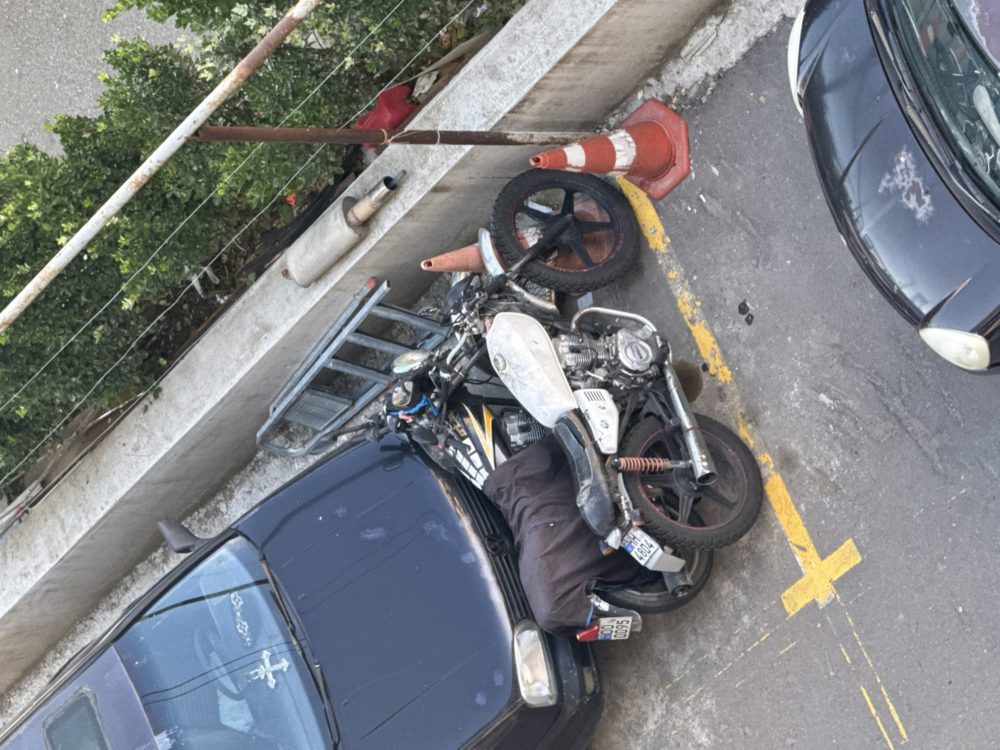 In Tbilisi, Georgia, a black car is parked next to a motorcycle. The motorcycle is covered with a black cloth and parked in front of the car, partially obscuring its front. A silver ladder is leaning against a concrete barrier behind the motorcycle. To the right of the motorcycle, there are two orange traffic cones, one of which has a white stripe. A rusty metal pole with wires attached stands near the cones. Behind the pole, there is dense green foliage. In the foreground, a yellow parking line is visible on the asphalt, along with what appears to be another car partially visible at the bottom of the frame. The scene suggests an outdoor parking area.