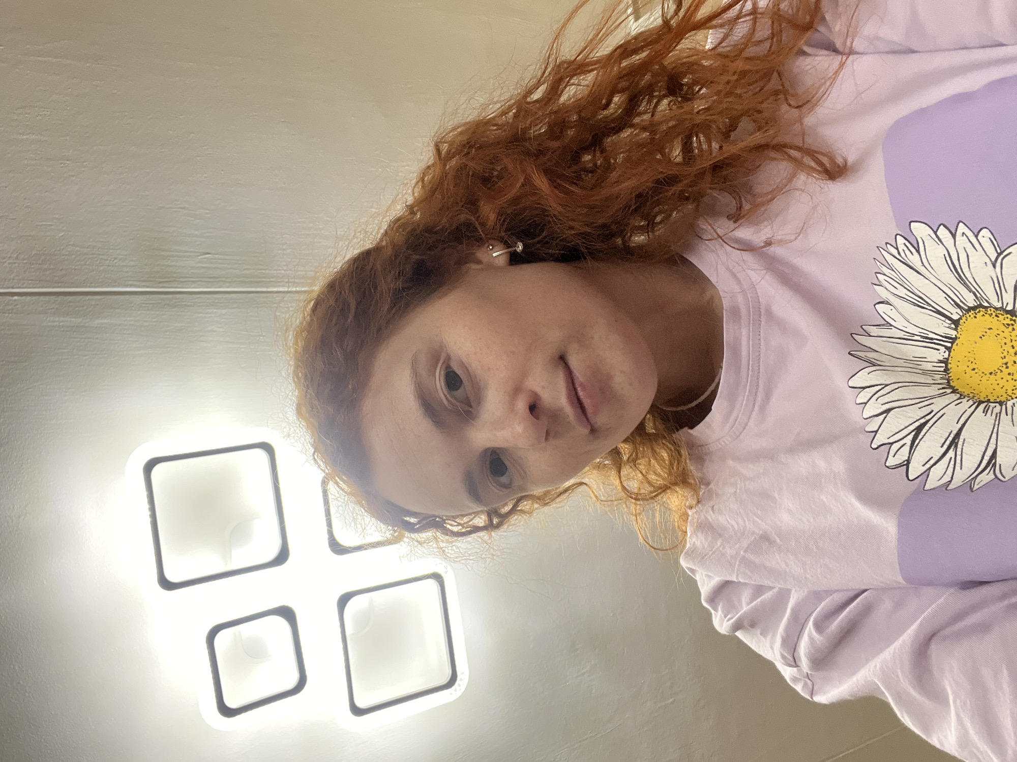 A young woman with red curly hair poses for a selfie. She is wearing a lavender t-shirt with a large daisy print. The background appears to be an interior, with a distinctive square-shaped ceiling light fixture visible. The image was taken in Chaikovsky, Russia.