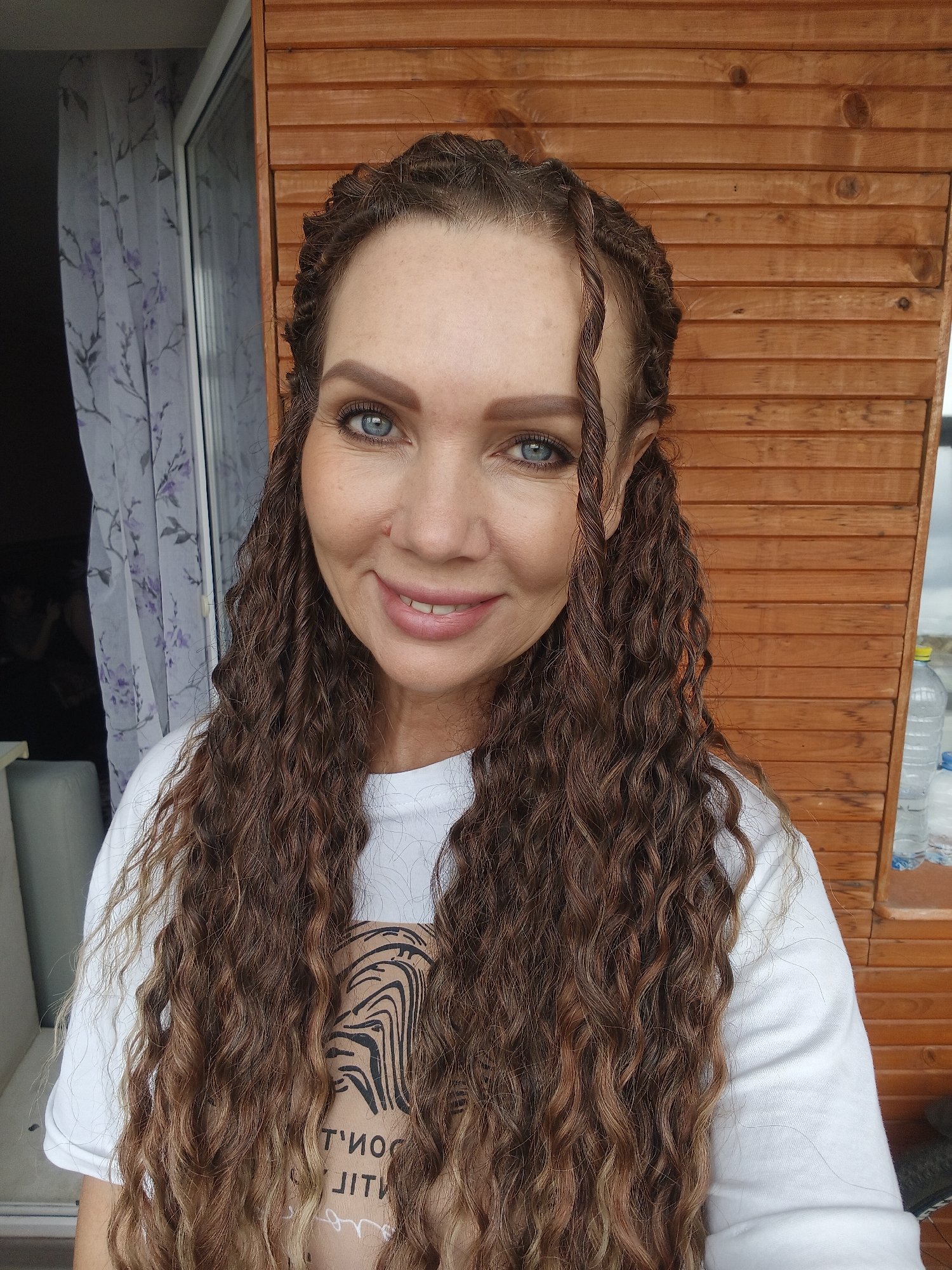 An adult female, positioned centrally, is depicted smiling directly at the viewer. Her long, heavily crimped or wavy hair, predominantly brown with lighter auburn or blonde highlights, extends past her shoulders. Two thin, twisted strands (rope twists or small braids) frame her face near the temples. She has prominent blue eyes and well-defined eyebrows. A small reddish mark is visible on her upper right cheek/nasal area. She is wearing a white short-sleeved t-shirt. Beneath her left arm, the top portion of a light brown garment with a black, wavy pattern (resembling zebra stripes) is visible, displaying inverted text 'T'NO' and 'JIT', suggesting 'DON'T QUIT'. The scene is set in the city of Unknown, Unknown.
To the left of the subject, a window is partially obscured by a sheer white curtain adorned with a light purple floral pattern. Through the window, a dark interior space with indistinct shapes, including what appears to be a light-colored cushion and a human silhouette, can be seen. To the right of the subject, a vertical wooden wall composed of horizontal planks with visible grain and knots serves as the background. On a narrow ledge along this wooden wall, two partially filled clear plastic water bottles are visible. The scene is illuminated by ambient daylight.