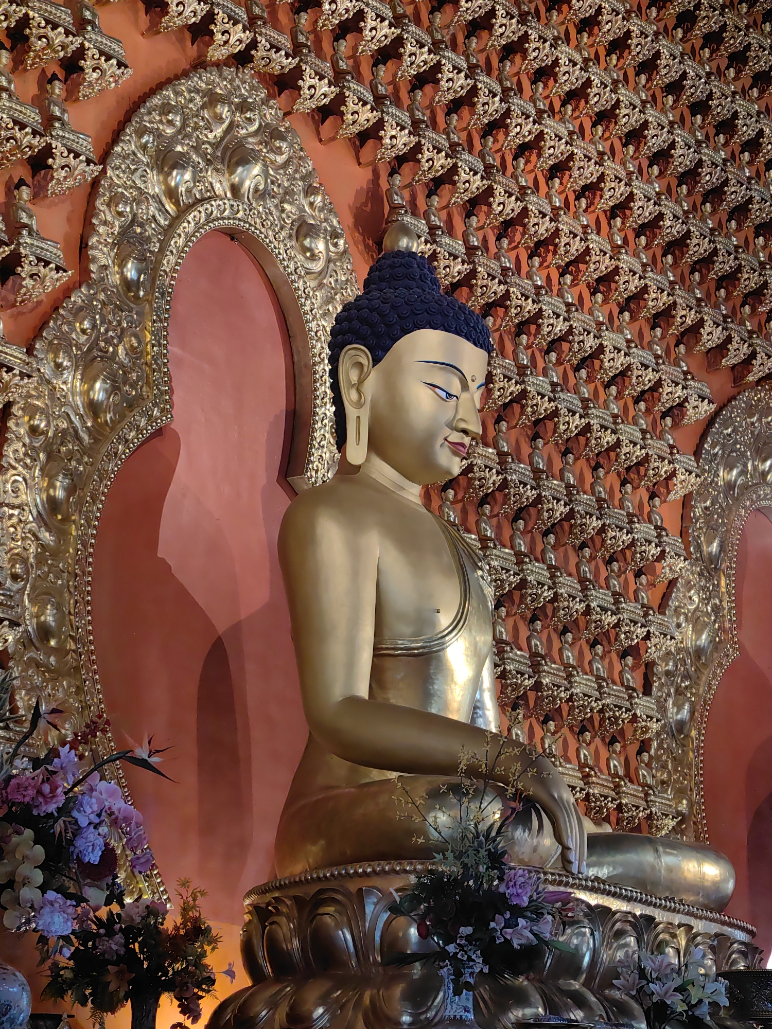 The photograph captures a close-up, side-profile view of a large, golden Buddha statue seated in a meditative pose. The statue is intricately decorated with ornate golden embellishments, particularly around its base and framing the Buddha's serene face. The background features a wall adorned with numerous smaller golden Buddha figures, each seated on a decorative bracket, creating a textured and visually rich tapestry. The wall itself appears to be a warm, reddish-orange hue. To the left and in front of the Buddha, a vibrant bouquet of pink and purple flowers adds a touch of color and natural element to the scene. The lighting highlights the metallic sheen of the gold and casts soft shadows, contributing to a sense of tranquility and reverence. The image does not provide information about a specific location within Parsa, Nepal, but rather focuses on the detailed depiction of a religious artifact and its surrounding artistic elements.