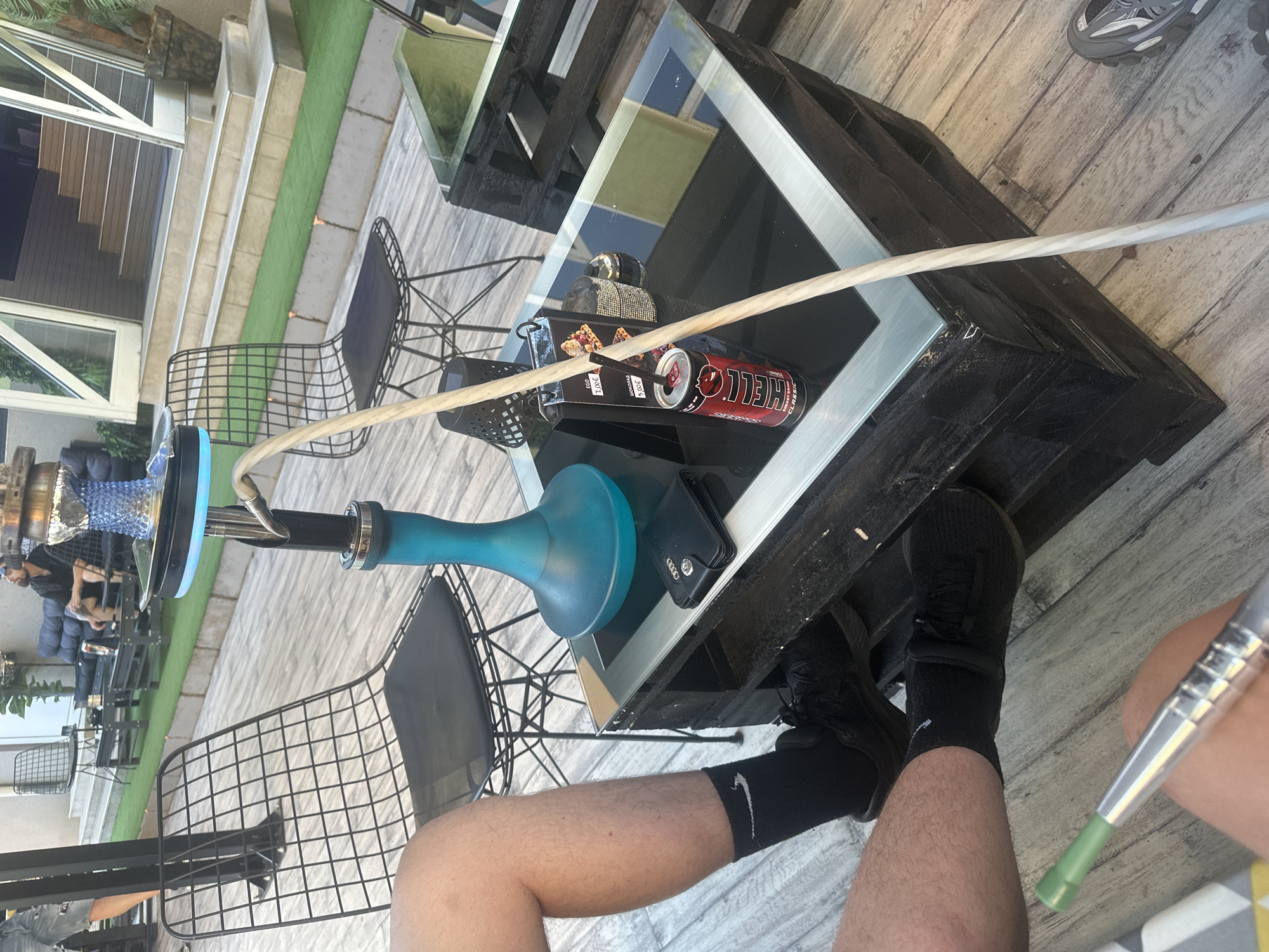 The image depicts an outdoor seating area, likely at a cafe or lounge. A black metal-framed chair with a black cushion is visible in the foreground, with a person's legs and feet wearing black socks and shoes to the left. A dark wooden table with a glass top sits in the center, displaying a blue hookah with an illuminated base, a wallet, and a red energy drink can. A light-colored hose extends from the hookah across the table. In the background, more seating areas with similar chairs and tables are arranged on a wooden deck and artificial grass. Stairs lead up to an entranceway with glass doors. Another person is seated in the background, wearing sunglasses and a dark shirt, looking upwards. The overall scene suggests a relaxed, casual atmosphere.