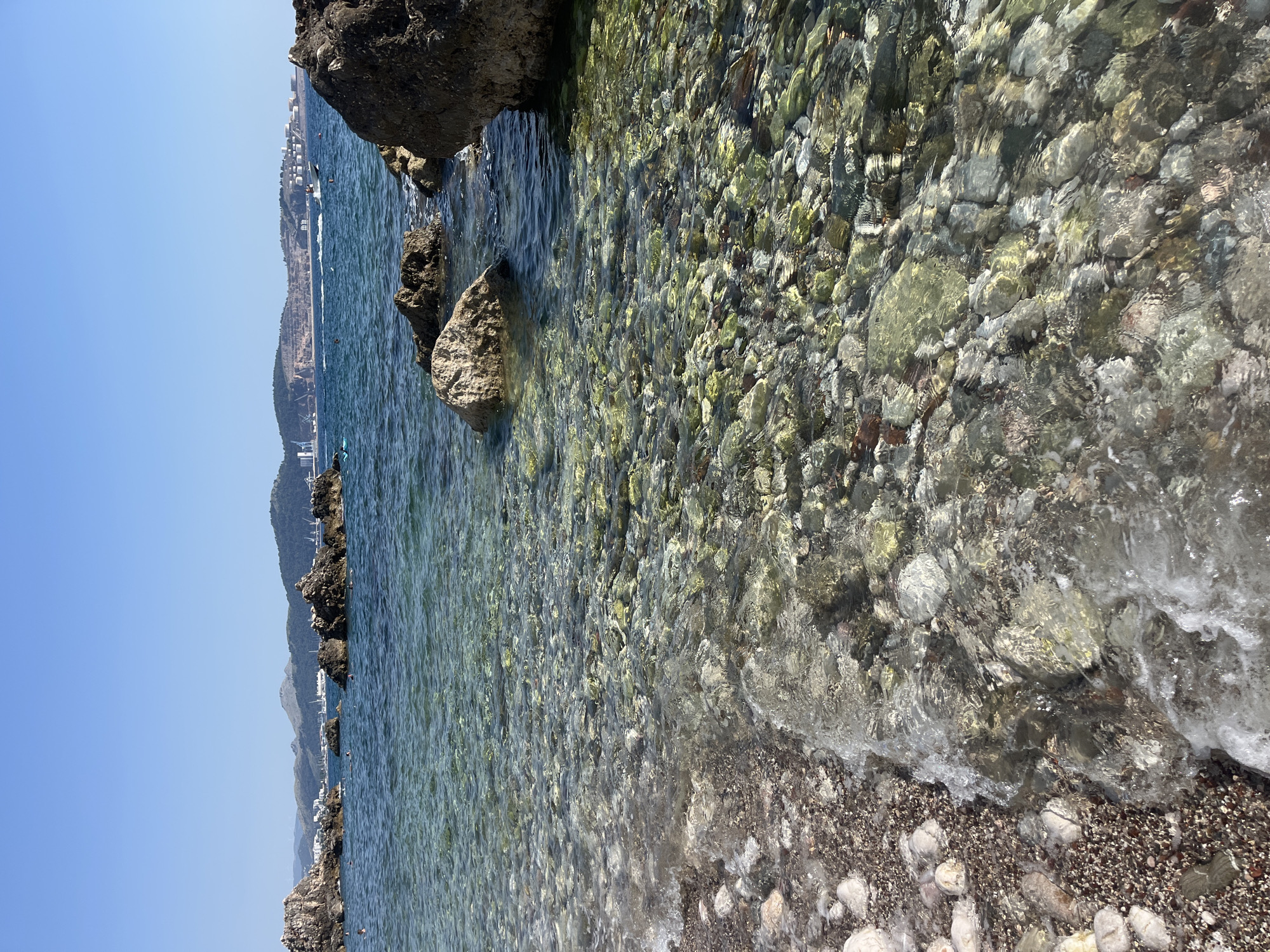 This is a photograph taken from a rocky shore, looking out towards the sea with a coastline in the distance. The water is clear, revealing a bed of colorful pebbles beneath the surface. Small waves are gently lapping at the shore, which is also composed of pebbles and rocks. In the background, a hilly coastline is visible, dotted with buildings and a road. The sky is a clear, bright blue, suggesting a sunny day.
The photograph captures a serene natural setting, likely a beach or cove. The clarity of the water and the abundance of smooth pebbles are prominent features. The distant coastline, with its vegetation and structures, hints at human habitation along the sea. The presence of people can be inferred from small figures visible in the water and along the shore in the background, suggesting that this is a popular spot for swimming or relaxation.
The lighting indicates it's daytime, likely mid-morning to late afternoon, given the bright sunlight and clear shadows. The overall mood is tranquil and picturesque, highlighting the beauty of the coastal landscape. There is no visible text in the image.