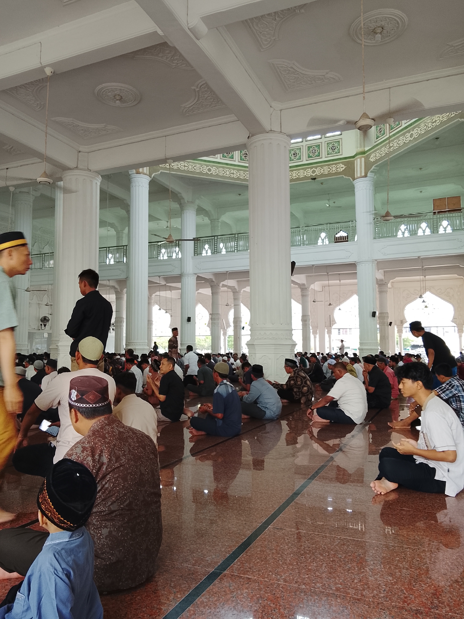 The image captures a gathering of people, likely men and boys, seated on the floor inside a spacious, well-lit mosque. The mosque features ornate white columns, intricate ceiling designs, and high arched windows. Natural light illuminates the scene, reflecting off the polished red floor. Many individuals are wearing prayer caps (peci) and traditional attire, suggesting they are participating in a religious activity, possibly congregational prayer. Some are looking towards the front, while others are engaged in quiet conversation or looking at their phones. Fans are visible on the ceiling, indicating a warm climate. The overall atmosphere appears to be one of reverence and community. The description of Lhokseumawe, Indonesia, pertains to the location of the mosque where this event is taking place.