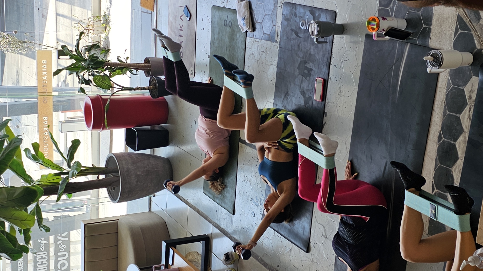 Inside the Baikastudio in Puebla, Mexico, four women engage in a leg workout. They are positioned head-down on yoga mats, their legs suspended in the air with resistance bands. Three of the women hold dumbbells in their hands. Various items, including water bottles and phones, are placed beside the mats. The room is decorated with several potted plants, a lounge chair, and a table. The studio’s name is visible in reverse on the glass wall.