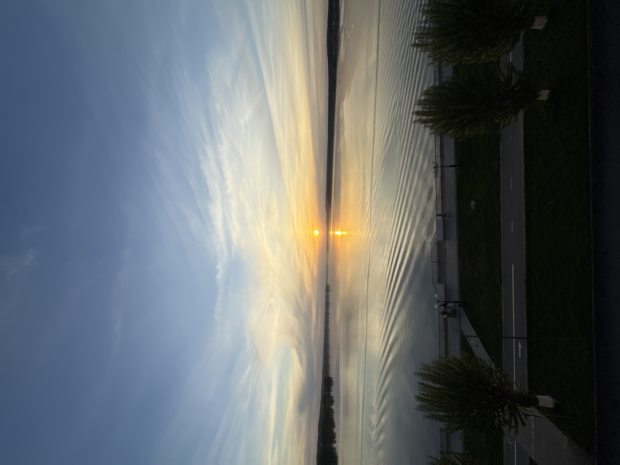 The image depicts a sunset over the Votkinsk Reservoir in Votkinsk, Russia. The sun is centered, reflecting intensely on the water's surface, creating bright glare. The sky above shows streaky clouds. The reservoir is framed by land masses on either side; the right side features a landscaped area with a path, trees, and a fence lining the water's edge. The path has painted lines, suggesting a pedestrian walkway. A couple of people are standing by the fence looking towards the water. The left side land masses are less visible, partially obscured by the bright light. There are distinct patterns of ripples across the water.