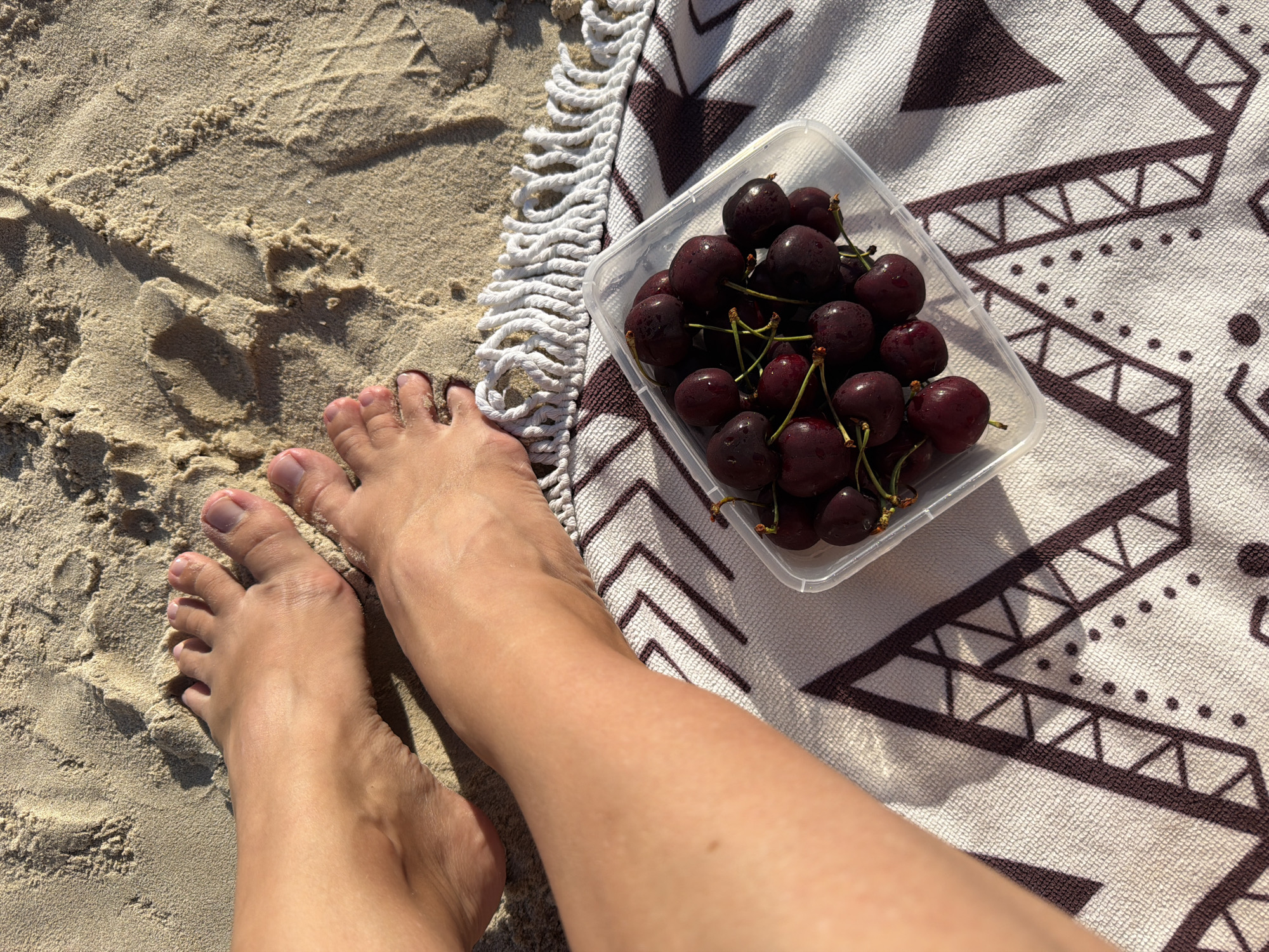 The image shows a pair of bare feet resting on a sandy beach. In front of the feet, on a patterned towel with fringe, is a clear plastic container filled with dark red cherries. The sand has some ripples and footprints. The lighting suggests it is daytime, with shadows cast by the feet and the cherries. The location is identified as the city of Bat Yam, Israel.