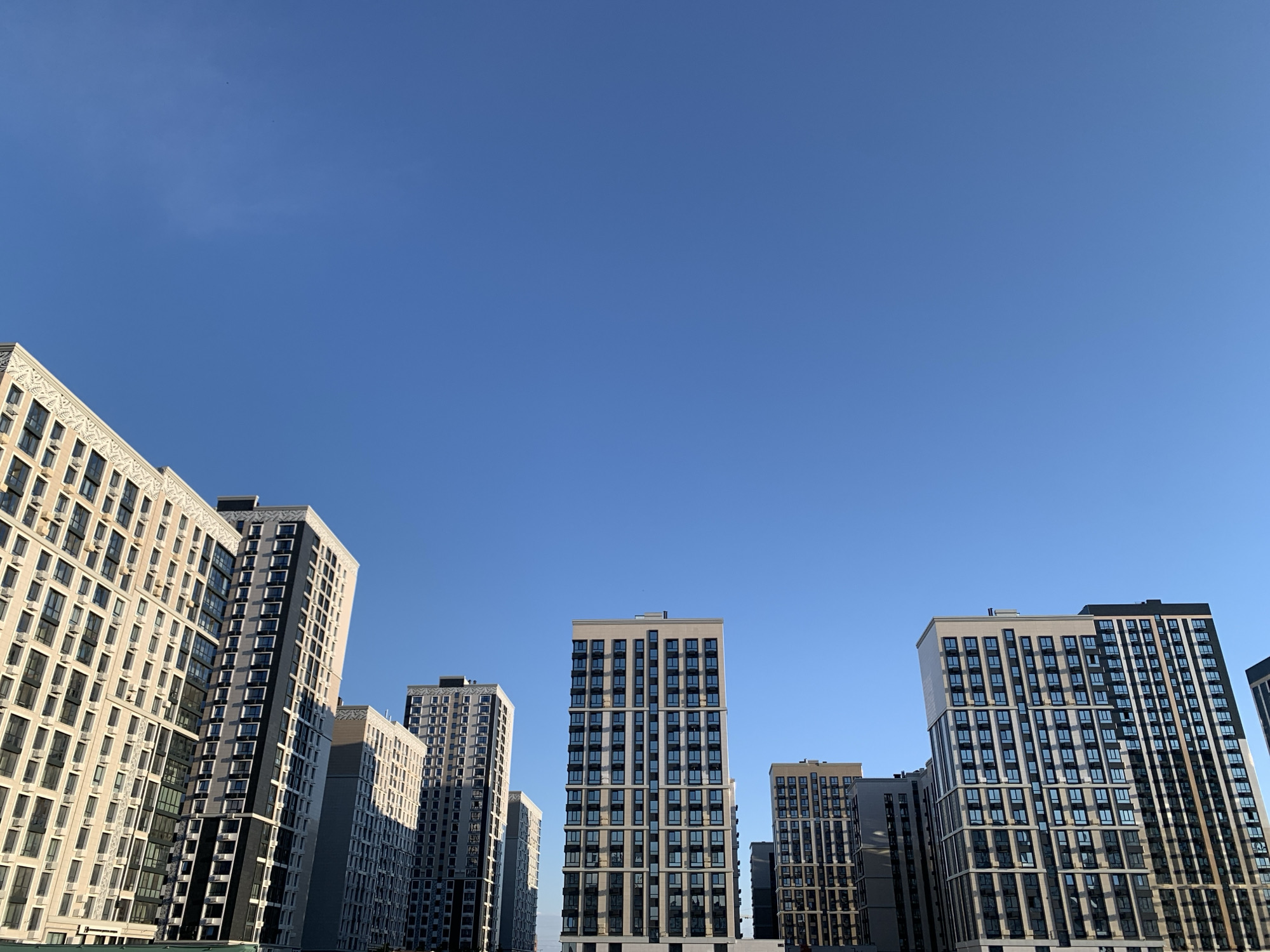 This media file captures a low-angle view of several modern, high-rise apartment buildings against a clear, bright blue sky. The buildings exhibit a variety of architectural styles, with some featuring light-colored facades and decorative patterns along the top edges, while others have darker, more geometric designs with numerous windows. Air conditioning units are visible on the exteriors of some buildings. The scene is bathed in sunlight, suggesting it is daytime, likely late morning or early afternoon, given the shadows cast. The clear sky indicates fair weather. The arrangement of the buildings suggests a residential development or a city district with contemporary architecture. There is no visible text in the image. The location context provided is Stavropol, Russia.