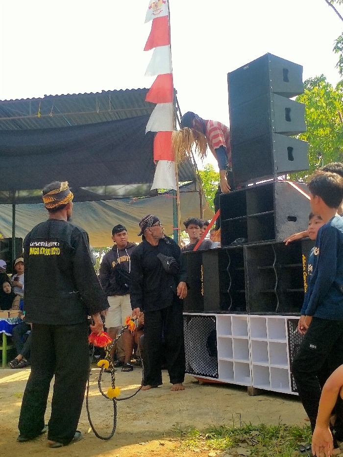 This outdoor scene in Plosorejo, Indonesia, depicts an informal event featuring a performance in front of a large sound system. In the foreground, two adult males in traditional black attire stand on dry, uneven ground. The man on the left, with his back mostly to the camera, wears a black shirt with white text, a yellow and brown braided headband, and holds a dark, braided rope adorned with yellow and orange tassels. The man in the center, barefoot, wears a black shirt, black pants, a patterned black and white headwrap, a dark fanny pack, and also holds the same tasseled rope.
To the right of the central figures, a tall stack of black modular speaker cabinets forms a substantial sound system, with the name "Abim Candras" visible on some units. White, cube-like enclosures are positioned at the base of the speakers. Elevated on top of the sound system, a person is dramatically bent forward, with long, light-colored material (resembling straw or hair) hanging from their head. This individual wears a red and white striped shirt over a blue long-sleeved shirt.
Behind the sound system, a bamboo pole flies several red and white rectangular flags. Numerous spectators, primarily young to middle-aged males, observe the scene from around the sound system. In the far left background, under a dark fabric canopy supported by poles, several people, including women, are seated. The ground is dry earth with sparse patches of green vegetation, illuminated by bright daylight.