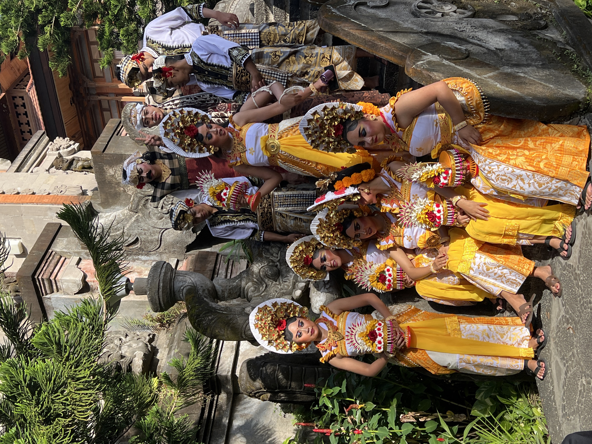 In Denpasar, Indonesia, a group of performers dressed in traditional Balinese attire poses for a photograph on stone steps. The performers, including men and women, are adorned in vibrant yellow and white costumes, elaborate headdresses, and intricate jewelry. Some hold decorative baskets filled with flowers. They are positioned on a tiered set of stone steps, with lush green foliage and ornate temple architecture in the background. The lighting suggests it is daytime, with sunlight casting shadows and highlighting the rich details of their outfits and the surrounding environment.