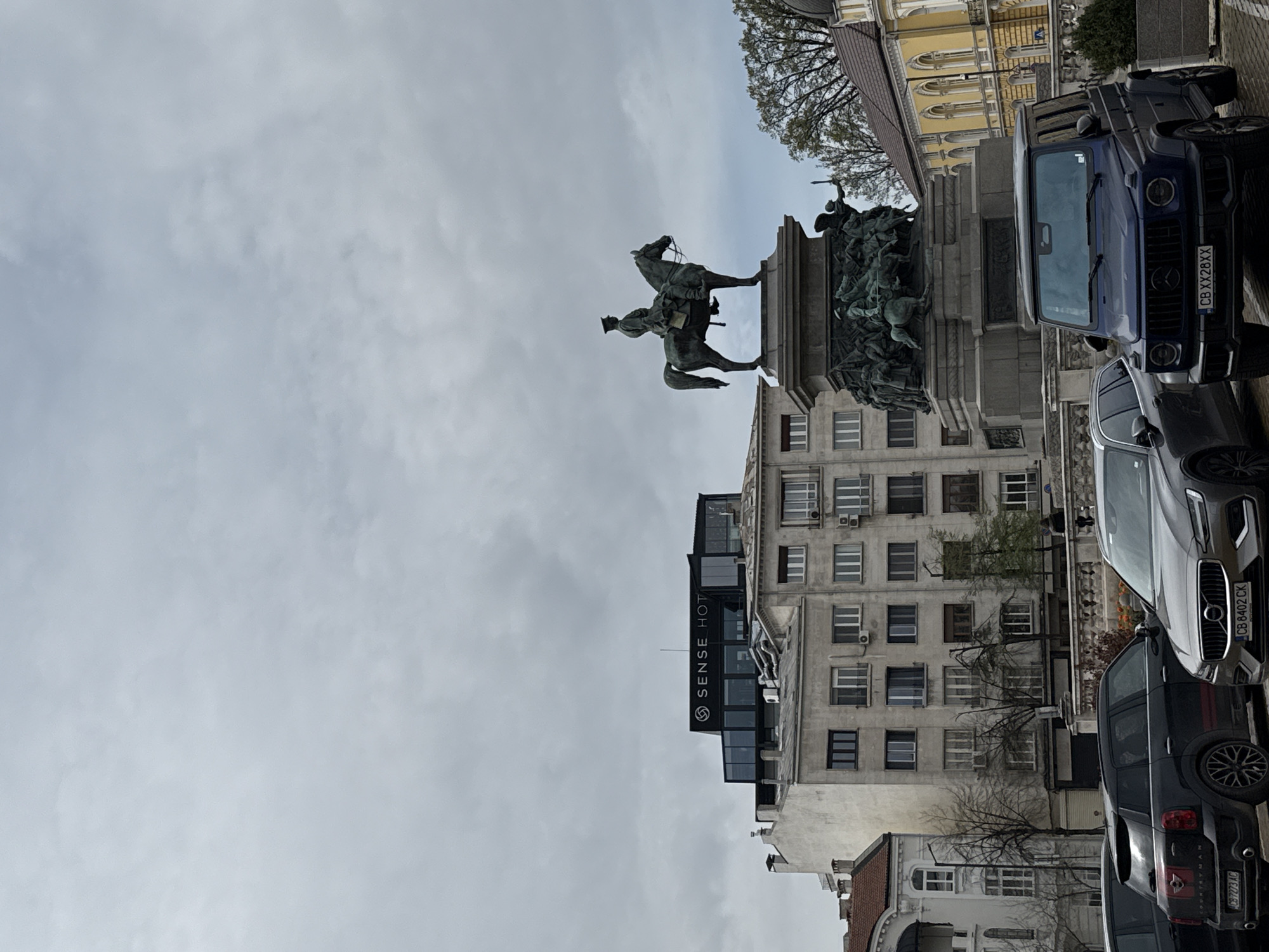 The image captures an urban street scene in Sofia, Bulgaria, centered around a large bronze equestrian statue, which is likely the Tsar Liberator Monument. The statue depicts a mounted figure, facing right, with an elaborate stone pedestal adorned with additional bronze figures and reliefs at its base.
On the right side of the frame, several cars are parked along the street. A prominent dark blue Mercedes G-Wagon is visible in the foreground, displaying a Bulgarian license plate "CB XX2BXX," with "CB" indicating Sofia. Behind it, a silver SUV, possibly a Volvo, bears the plate "CB 8402 CK," and a dark Mini Cooper Countryman shows "CB 7273 AC."
Various multi-story buildings form the backdrop. One modern building features "SENSE HOTEL" written vertically on its dark facade. Other older buildings with numerous windows and visible air conditioning units line the street. Bare trees and some with light green leaves are scattered throughout the scene. The sky is largely overcast and light grey, suggesting a cloudy day. There are no specific activities or people highlighted, presenting a static street view of a prominent monument and its urban surroundings.