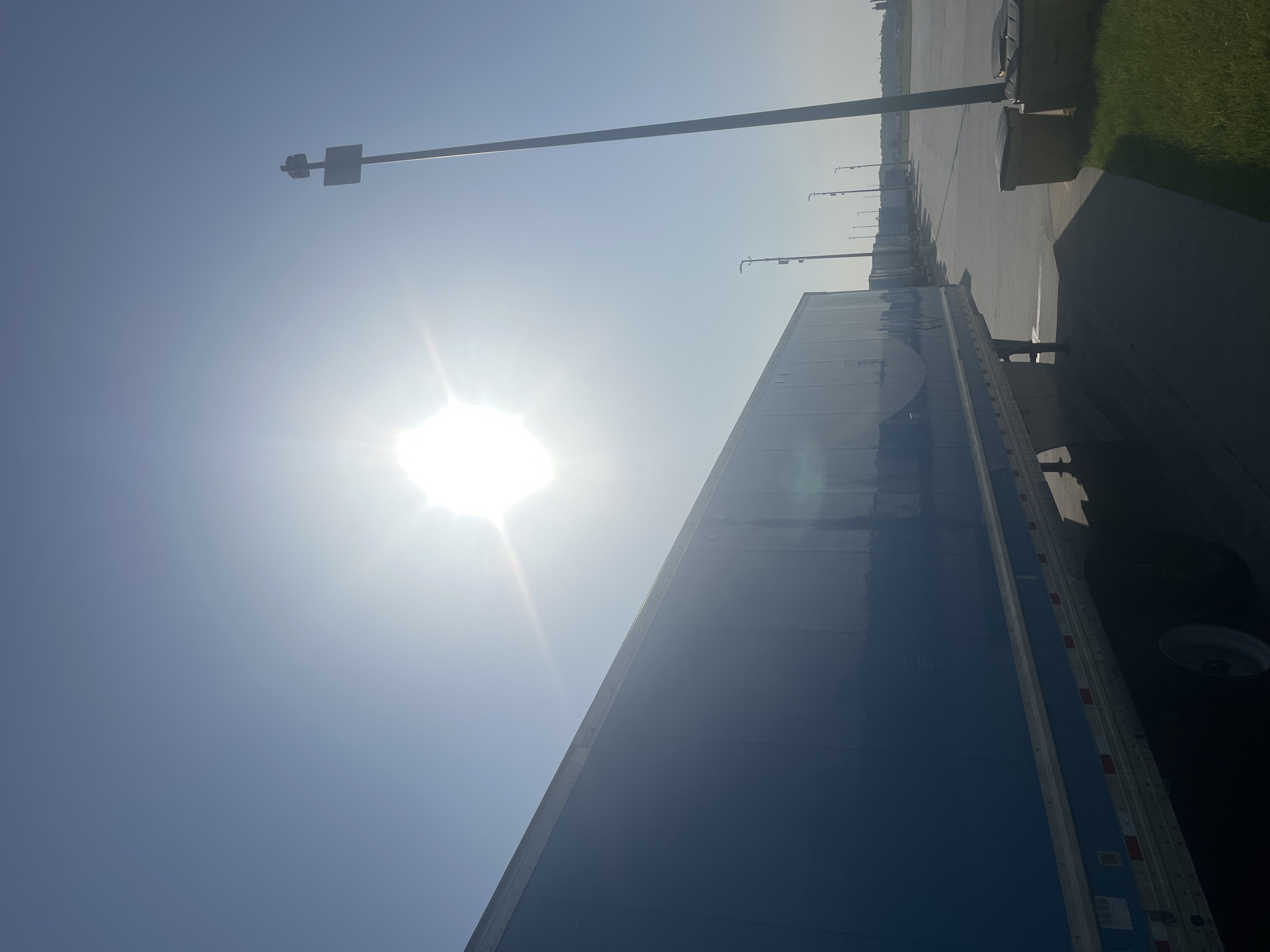 The image shows a bright, sunny day with the sun high in the sky, causing lens flare. In the foreground, the side of a blue semi-trailer is visible, angled towards the right of the frame. The trailer is parked in what appears to be a large lot, possibly a truck stop or distribution center. Several other semi-trailers are parked in a line behind the first one, receding into the distance. A tall pole with a sign and what looks like a siren or speaker is visible on the right side of the image. Further to the right, two large yellow trash bins are situated on the pavement next to a grassy area. The overall environment suggests an industrial or logistical setting. This scene is likely located in Pooler, Georgia, United States, which is known for its significant trucking and logistics presence.
