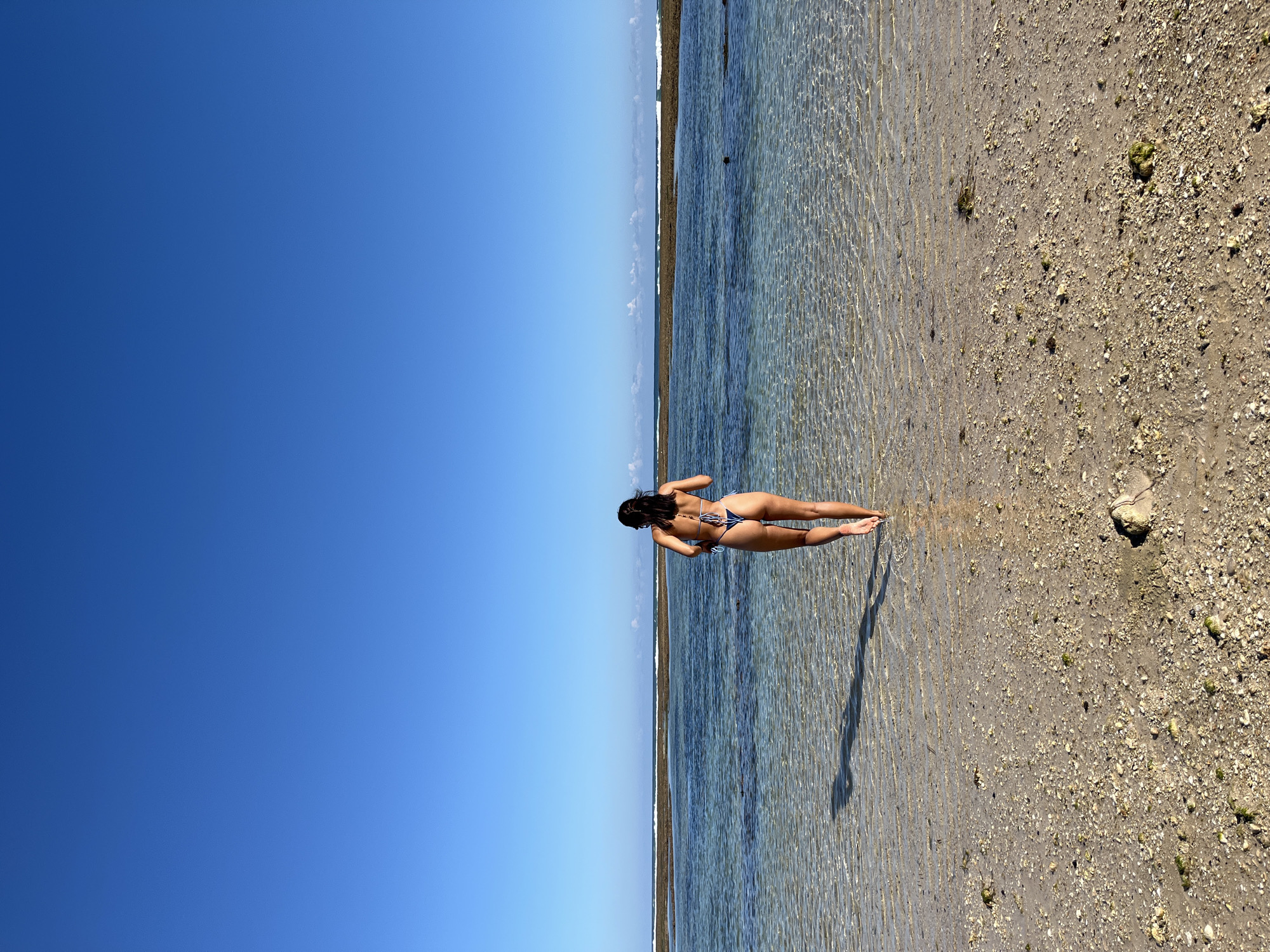 The image captures a serene coastal scene in Kuta Selatan, Indonesia, featuring a lone woman standing in very shallow, clear water. The woman, viewed from behind, is wearing a dark blue or black two-piece bikini with a white or light blue pattern. She stands with her body slightly turned, looking out towards the distant horizon where the sky meets the water.
The setting appears to be a beach or a low-tide area, characterized by exceptionally clear, calm water that reveals the sandy, somewhat rocky seabed beneath. Gentle ripples are visible on the water's surface, indicating minimal movement. To the right, the shore consists of light-colored sand mixed with small pebbles and sparse patches of green marine growth. The sky above is a brilliant, cloudless blue, suggesting perfect, sunny weather.
The scene is brightly lit by what appears to be midday or early afternoon sunlight, casting a distinct shadow of the woman onto the water and sand. This lighting emphasizes the clarity of the water and the textures of the shore. The overall impression is one of peacefulness and natural beauty, with the woman enjoying the tranquil coastal environment. There is no visible text in the image.