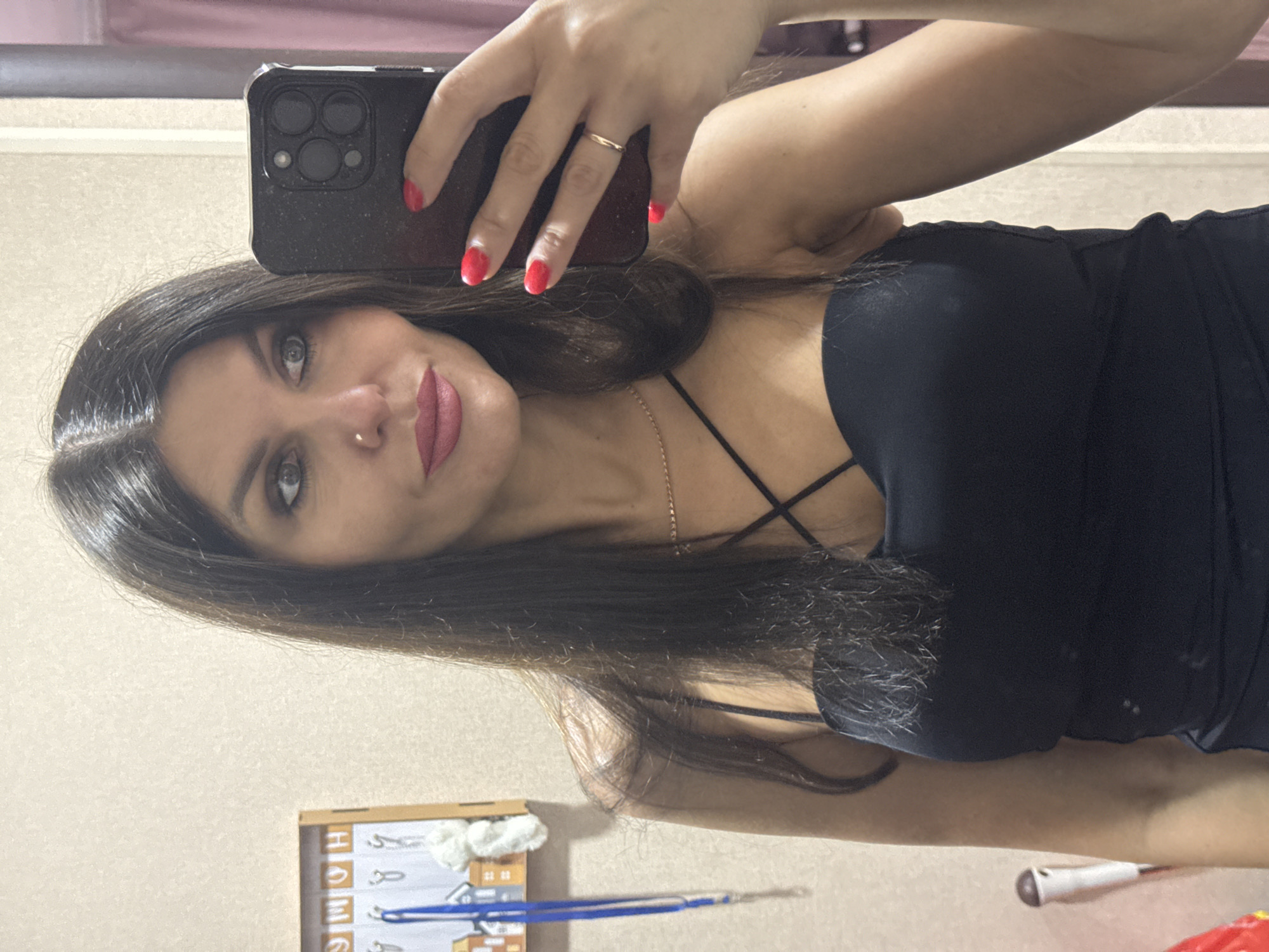 A woman with long, dark hair and makeup is taking a selfie in a mirror. She is wearing a black, strappy dress and has bright red nail polish on her fingers, one of which is adorned with a thin gold ring. The background appears to be a wall with a framed object containing the word "HOME" and hooks, possibly a key holder. The image is captured in Chaikovsky, Russia, based on the metadata.