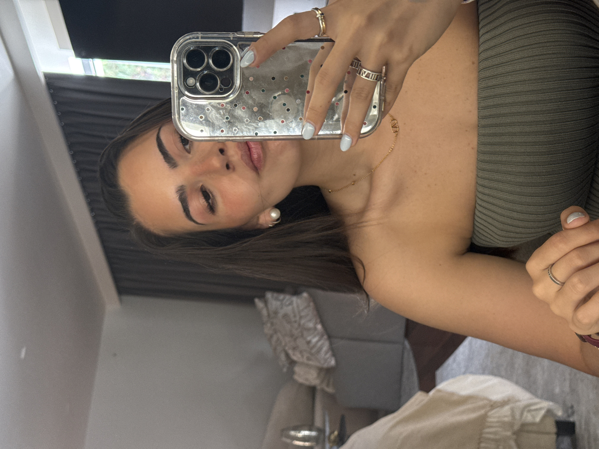 A young woman is taking a selfie in what appears to be an interior space. She is holding a smartphone with a clear, glitter-speckled case, and the reflection shows part of her face, including her eyes, eyebrows, lips, and a pearl earring. She is wearing rings on her fingers and a delicate gold necklace. She is dressed in what seems to be a ribbed, strapless top, possibly olive green in color. In the background, elements of a room can be seen, including what looks like a pillow with a pattern, a portion of a chair or sofa, a window, and some curtains. The location in the city of San Bernardino Tlaxcalancingo, Mexico, is not discernible from the image.