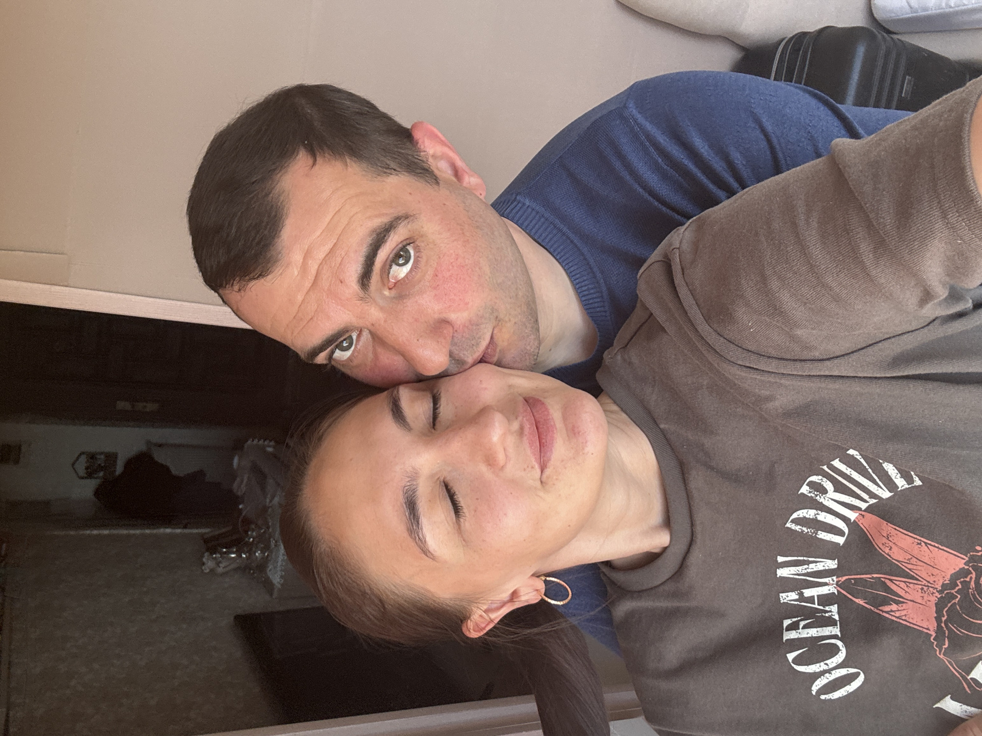 An intimate indoor scene captures a man kissing or nuzzling a woman's cheek. The image, taken from an overhead perspective and rotated 90 degrees clockwise, shows both individuals lying down.
The man, with dark hair, green/hazel eyes, and light stubble, wears a blue long-sleeved sweater. His face exhibits some redness, possibly indicating a blush or natural skin tone. He is positioned closely with the woman, directing an affectionate gesture towards her.
The woman, with dark hair pulled back and wearing a gold hoop earring, has her eyes closed and a soft, content expression, suggesting comfort and happiness. She is wearing a dark gray or brown t-shirt. Visible text on her shirt reads "OCEAN DRIVE," alongside a stylized design that appears to be an insect or moth.
In the blurred background, a black suitcase is visible, along with other indistinct elements of what appears to be a room. The overall atmosphere conveys tenderness, intimacy, and affection between the two subjects, possibly in Sochi, Russia.