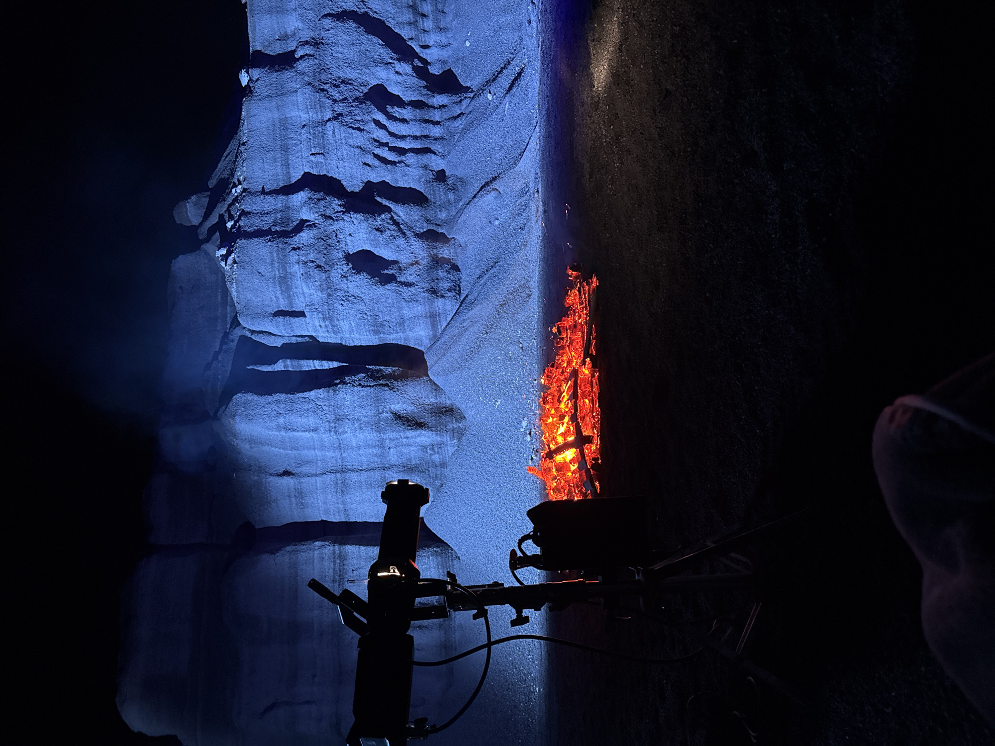 The image depicts a volcanic cave in Teguise, Spain. The left side of the cave wall is illuminated in a blue hue, revealing layered rock formations. To the right, a vertical seam in the rock face is lit by a bright, orange-red light, resembling a fire. A dark silhouette of what appears to be a lighting rig or camera equipment is positioned in the foreground. The right side of the image is obscured in shadow, with a hint of what may be a person’s hand or arm visible in the lower right corner.
