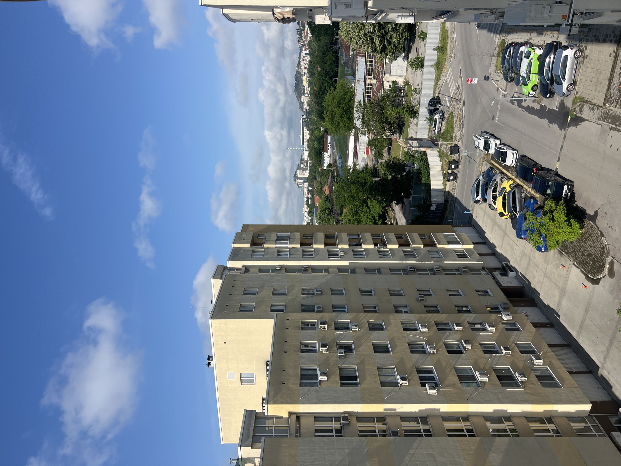 This high-angle daytime photograph captures an urban scene in Plovdiv, Bulgaria, looking down from an elevated perspective. On the left side of the frame, a tall, multi-story building, likely residential or commercial, is the dominant foreground subject. Its facade is a light beige or yellow, featuring numerous dark-paned windows and many external air conditioning units mounted beneath them.
To the right, a paved street and parking area are visible. A diverse collection of compact cars in various colors—including white, black, light green, yellow, and blue—are parked along the curb and in a designated lot. A distinctive detail is a white car that appears overturned or on its side on a small patch of grass adjacent to the road. A red "STOP" sign is clearly visible on a pole near the parked vehicles.
Beyond the immediate street, the mid-ground and background reveal a sprawling cityscape. Abundant green trees are interspersed with various buildings, including what appears to be an industrial structure with large windows and a tall, slender chimney-like feature in the distance. Rolling hills or mountains covered in vegetation form the furthest backdrop.
The sky, visible in the upper left quadrant, is bright blue with scattered fluffy white cumulus clouds, indicating clear and sunny weather. Bright sunlight casts distinct shadows, suggesting it is mid-morning or afternoon. No people are discernible, and the primary activity observed is stationary parking, with the overturned car being a notable detail of a possible past event.