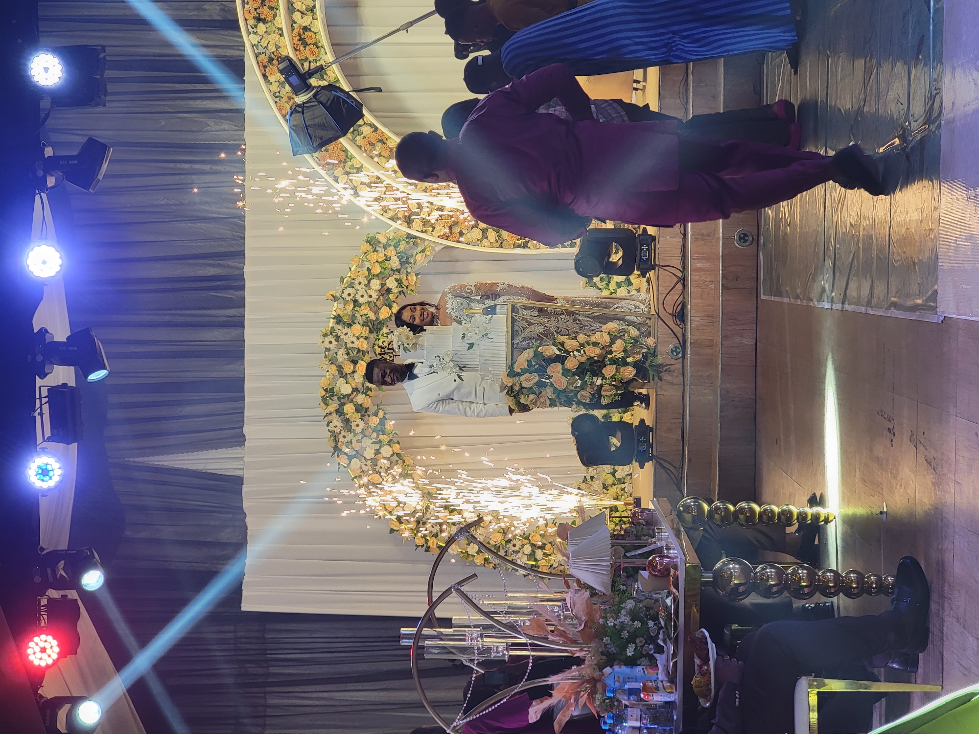 The image captures a vibrant indoor celebration, likely a wedding reception, filled with celebratory lighting and special effects. The scene is dominated by a newlywed couple standing prominently at the center. The bride, wearing a sparkling, intricately designed gown, beams with a joyful smile. The groom, dressed in a crisp white suit, stands beside her, looking towards the viewer. They are positioned in front of a grand, semi-circular floral arch adorned with white, cream, and peach-colored flowers. In front of them, a multi-tiered white wedding cake is visible, and dazzling pyrotechnics erupt from the ground, creating a shower of golden sparks that illuminate the couple and the floral backdrop.
The event space is theatrically lit, with multiple professional spotlights casting beams of blue, red, and white light from the left side of the frame. One spotlight clearly shows the text "MINI 270." A black softbox on a stand is also visible, indicating a professional photography setup. Several attendees are present; one individual in a purple garment stands near the couple with their back to the camera, while others, possibly guests or event staff, are visible in the background and foreground, some with cameras.
The backdrop features draped white and dark fabric, suggesting an elaborately decorated venue. In the foreground, a decorative golden circular structure and partial views of tables with refreshments add to the festive atmosphere. The artificial lighting and special effects confirm an evening event taking place in Barden Yana, Nigeria, emphasizing a moment of joy and grandeur.