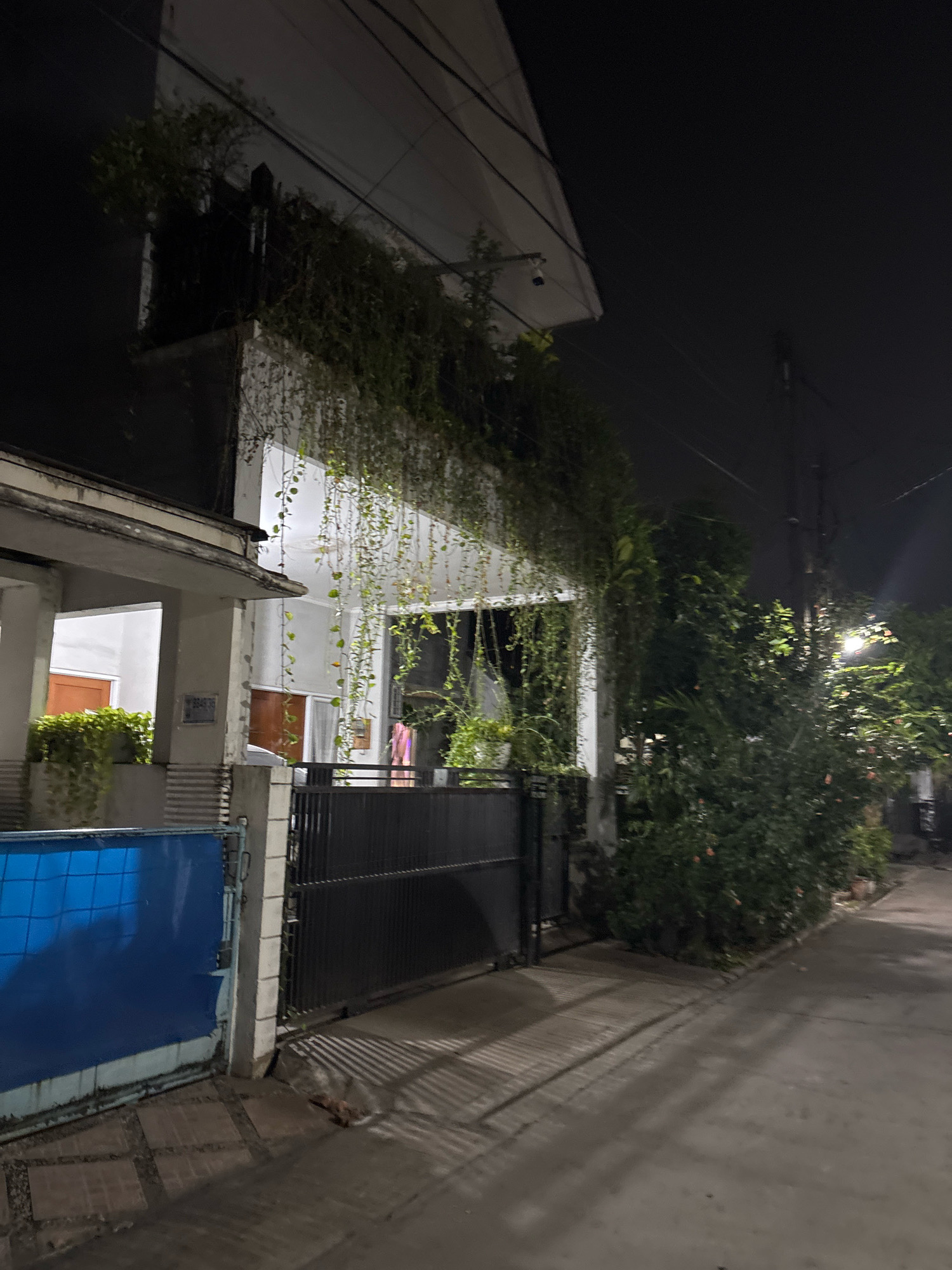 A nighttime outdoor shot shows a house in Bekasi, Indonesia. The house is multi-story and dark, with lush green vines cascading down its facade. A black metal gate stands in front of the property, partially obscuring a glimpse of the interior. To the left of the gate, a blue metal fence is visible, bordering a paved walkway that leads to the house. The street in front of the house is illuminated by a single streetlamp, casting shadows on the ground.
