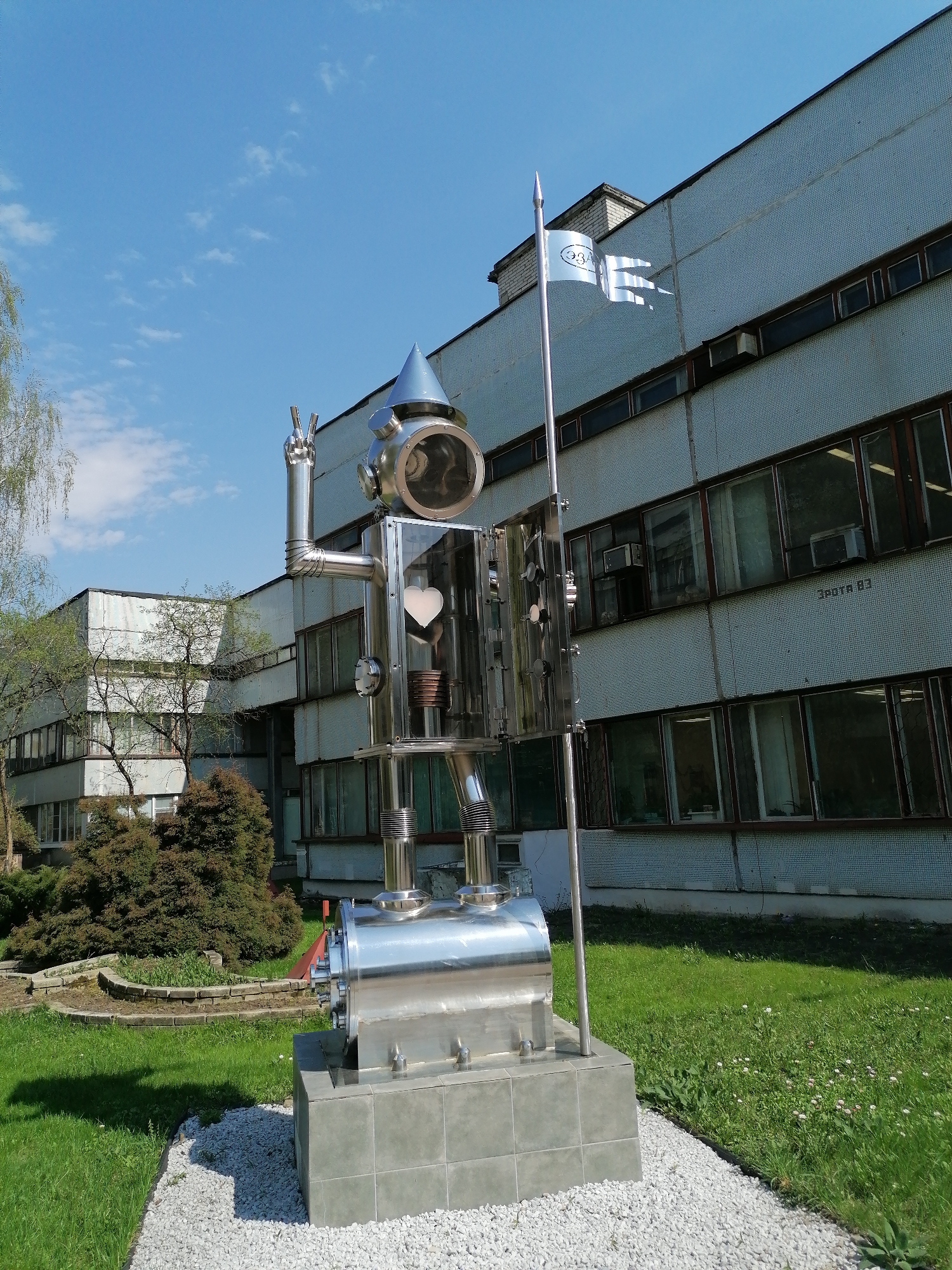 A stainless steel sculpture of a robot stands on a gray tiled platform, surrounded by white gravel, in a grassy area near a large, multi-story building. The robot has a round head with a conical top and a white heart visible in its chest area. One arm is raised with two fingers extended in a peace sign. A silver flag bearing a logo flutters from a pole attached to the robot. The building has a tiled facade and numerous windows. A small, manicured garden with bushes is visible to the left of the sculpture. The location is in the city of Unknown, Unknown.