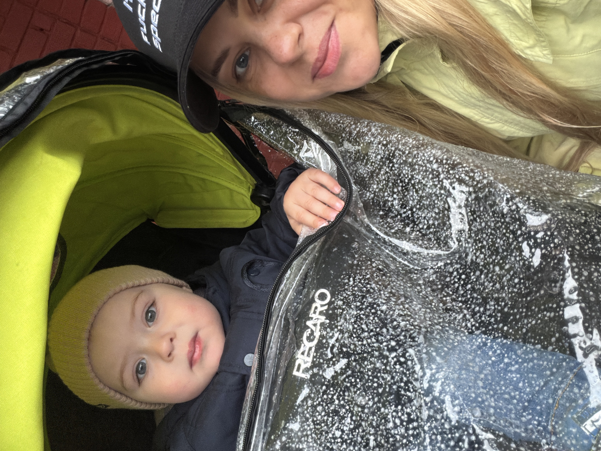 The image depicts an adult female and a young child in a selfie-style photograph taken in Votkinsk, Russia. The adult female occupies the right side of the frame, presenting with blonde hair visible beneath a black baseball cap. The cap features white fragmented text reading "It," "fuck," and "spec" on its front. She wears a light green collared jacket and has blue eyes, appearing to look directly at the camera with a slight smile.
To the left of the adult is a young child, likely an infant or toddler, seated in a stroller. The stroller's interior canopy is a vibrant lime green. The child wears a light beige knit beanie and a dark blue padded jacket, with blue jeans visible through a transparent plastic rain cover. The child has blue eyes and appears to be looking towards the camera.
The rain cover, prominently displaying the white "RECARO" logo on its dark fabric section, is covered in numerous small water droplets, indicating precipitation. A small portion of the child's left hand rests on the rain cover. In the background, a red brick wall is partially visible in the upper right corner, behind the adult's head and the stroller's canopy. The area directly behind the child in the stroller appears dark and obscured.