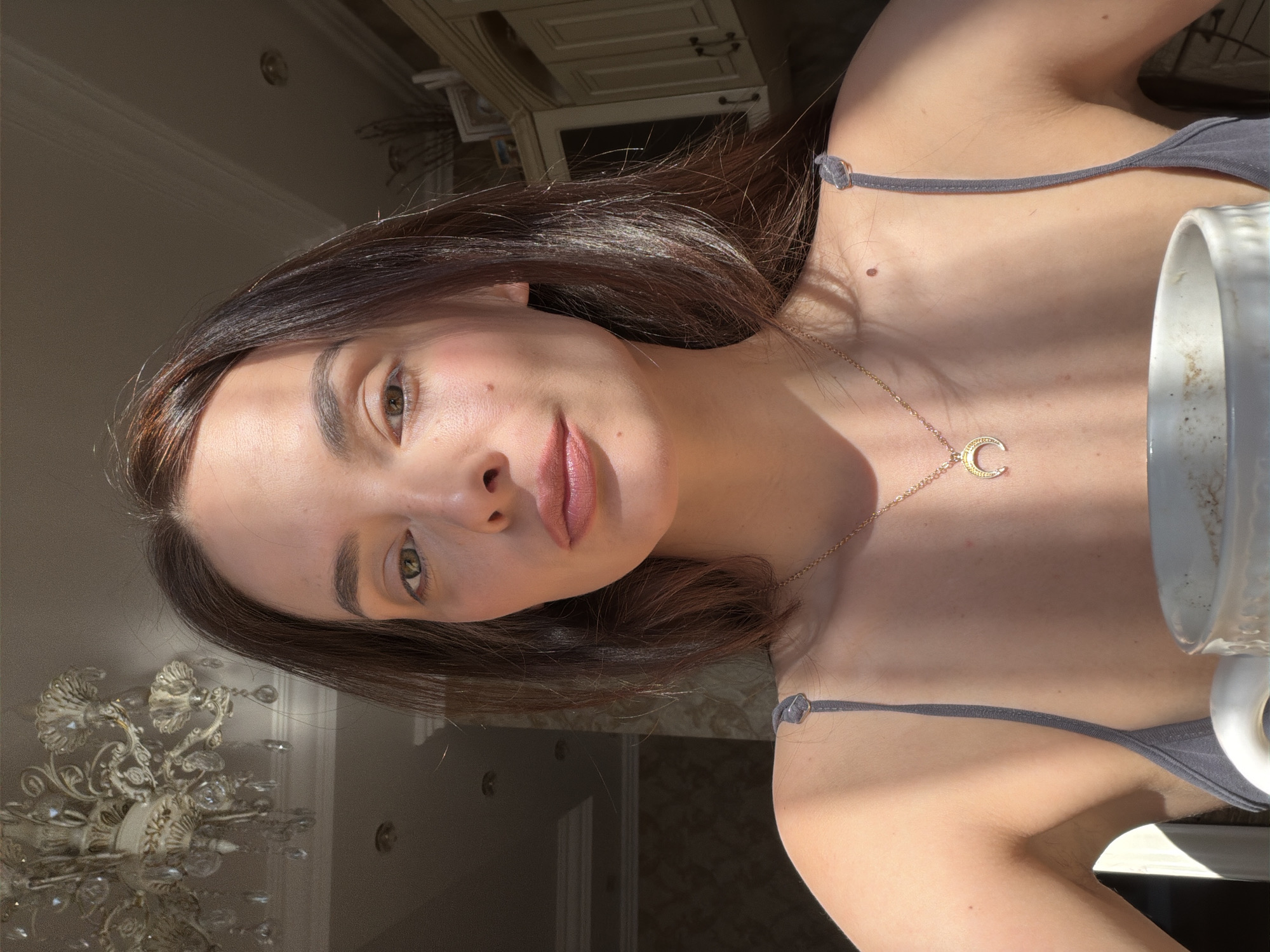 A woman with brown hair is looking forward, with the sun casting shadows across her face. She is wearing a gray tank top and a gold necklace with a crescent moon pendant. In the foreground, a white mug is partially visible. The background shows a chandelier and kitchen cabinets. The scene appears to be indoors in a domestic setting.