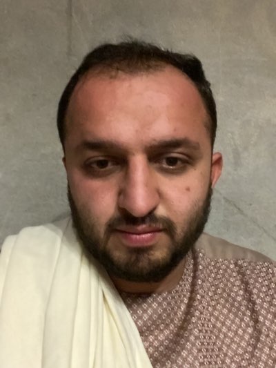 A close-up shot depicts a man with dark hair and a beard. He wears a patterned shirt and a cream-colored shawl draped over his shoulder. The background is a plain gray wall. The image's location cannot be determined as Kandahar, Afghanistan.