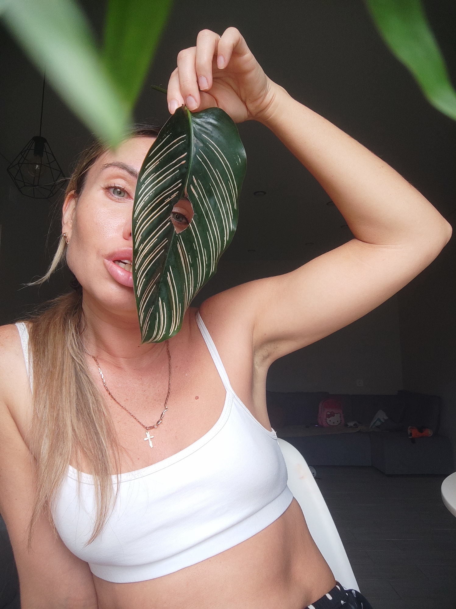 A woman in a white crop top holds a green leaf with white stripes in front of her face, partially obscuring it. She has blonde hair pulled back in a ponytail, a necklace with a cross pendant, and is looking at the camera. The background is a dimly lit interior of a room. A section of a gray couch is visible along with a pink backpack, some toys, and a white table. A hanging light fixture can also be seen. There is no evidence suggesting the location is in Votkinsk, Russia.