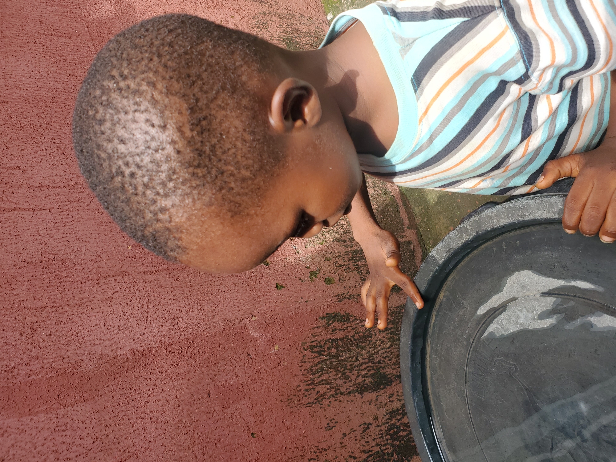 The media file captures a high-angle, close-up shot of a young child's head and upper torso, viewed from above and tilted to the side. The child has closely cropped dark hair and is wearing a striped t-shirt with alternating bands of light blue, white, dark gray, and orange. The child's head is resting on a textured reddish-brown surface, possibly concrete or painted ground, which appears to be outdoors due to the dappled sunlight and shadows. To the right of the child, a large, dark gray plastic basin or container is visible, partially filled with water that reflects the light and surroundings. The child's right hand is reaching towards the basin, with fingers slightly spread, suggesting interaction or curiosity. The overall scene appears to be a casual outdoor setting, possibly in a residential area, given the combination of the ground and the basin. The lighting suggests it is daytime, likely with bright sunlight casting shadows. There is no visible text in the image. The child's expression is not clearly discernible due to the angle and focus, but the posture and hand gesture might indicate engagement with the water or the container. The context provided indicates the location is Jos, Nigeria.