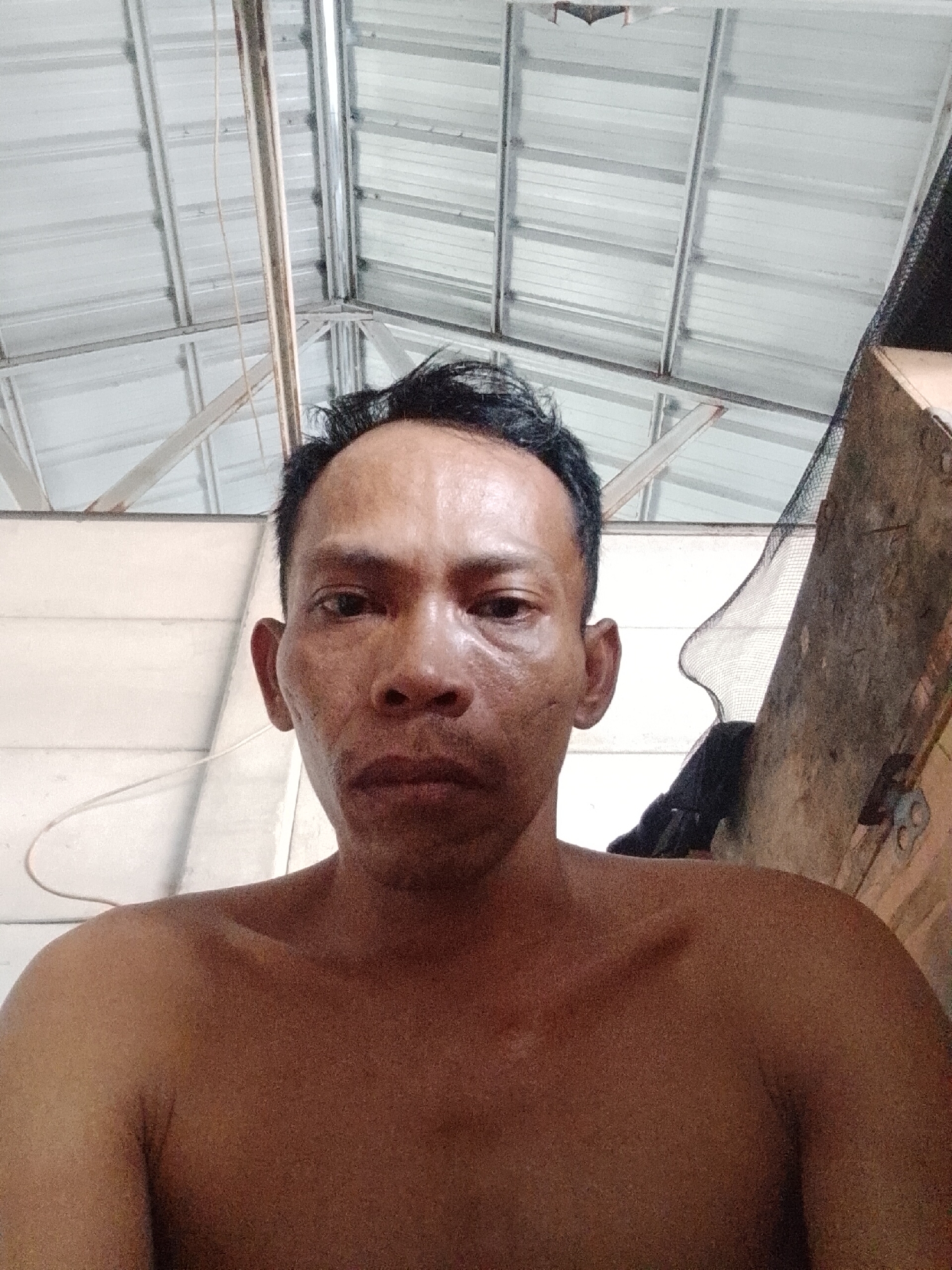 A medium close-up shows a shirtless man with dark hair indoors under a metal roof structure with white beams. The man's face has apparent uneven skin tone, and his expression is neutral. A wire or cord is visible near his left shoulder. To the right, there is a rough, possibly wooden wall with a dark, indiscernible object. The image was taken at a close distance, and the location is Bukit Ketok, Indonesia.