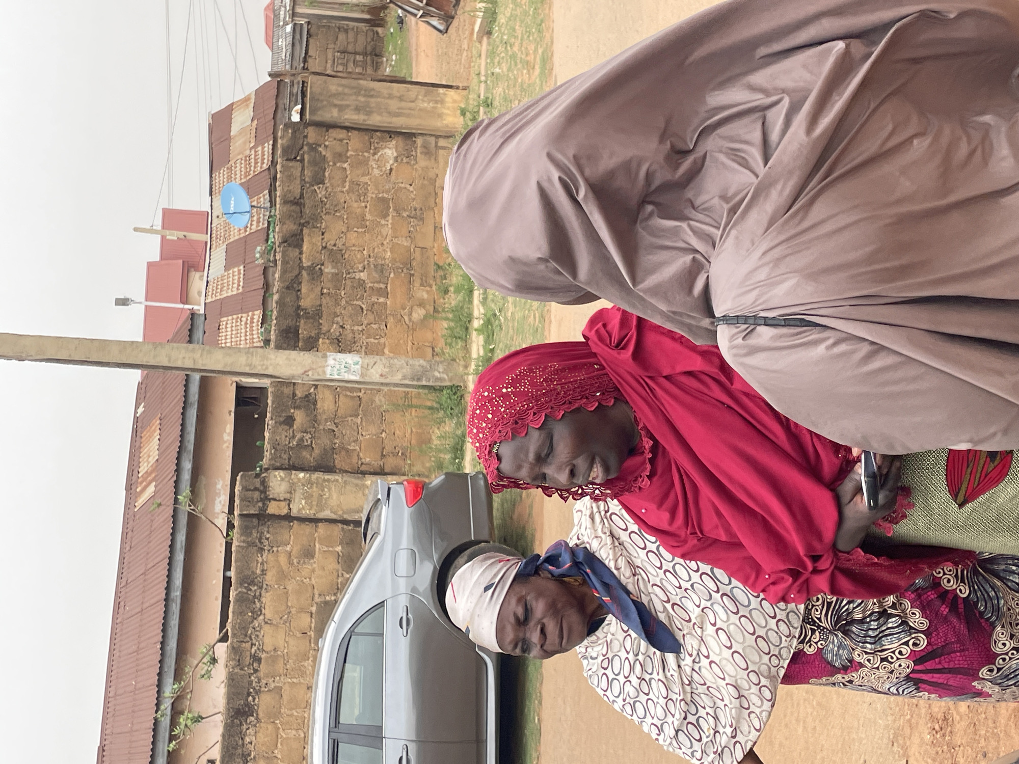 The image captures three women in an outdoor setting. The woman in the foreground wears a white and patterned top with a blue scarf. Next to her, a woman wears a red head covering and holds a smartphone. A third woman is partially visible, wearing a tan colored garment that covers her head and body. A light-colored car is parked nearby. The background includes buildings with brick and corrugated metal walls and roofs, and a blue satellite dish is visible. Based on the prompt's instructions, the location is the city of Jos, Nigeria.