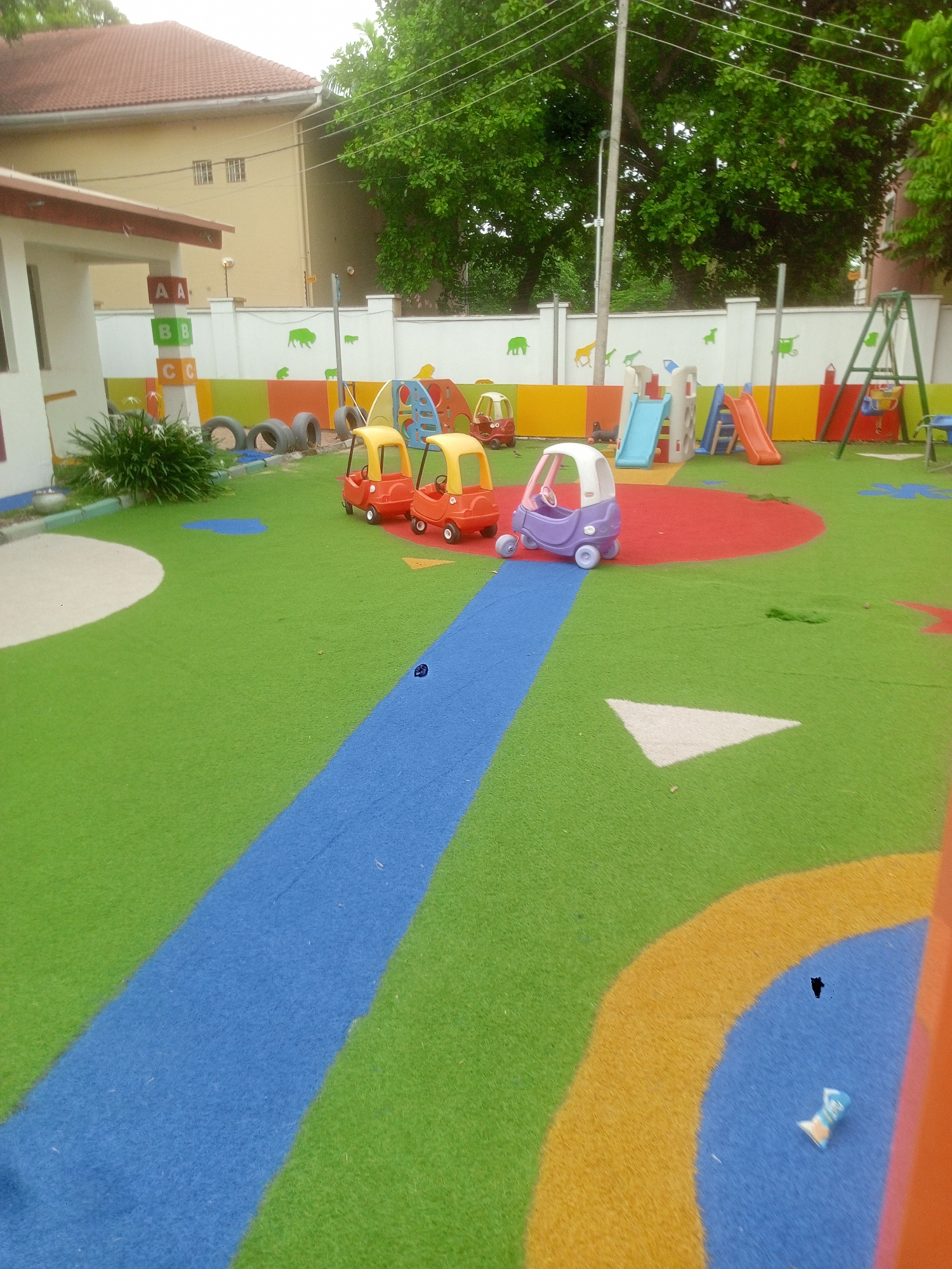 The image shows a brightly colored children's playground, likely located in Abuja, Nigeria given the architecture in the background. The playground has artificial grass with various colored shapes and pathways, including a blue "road" and a red circular area. There are four small plastic toy cars parked near the red area; three are red and one is purple. The playground is equipped with slides, a swing set, and other play structures. The perimeter is defined by a low wall decorated with animal silhouettes. There is also what appears to be a tower of colorful blocks with letters of the alphabet at the edge of the play area. In the background is a building with a brown roof.
