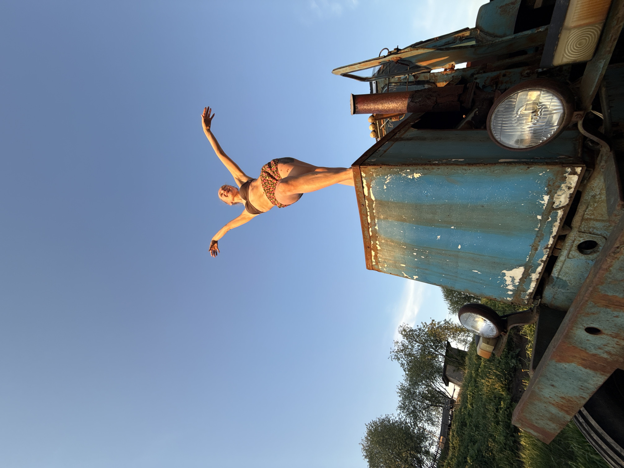 A woman in a bikini is diving off the hood of a weathered, blue and rusty truck. The background features a clear blue sky above and trees and a building below. The photograph was taken in Nizhnyaya Chermoda, Russia.