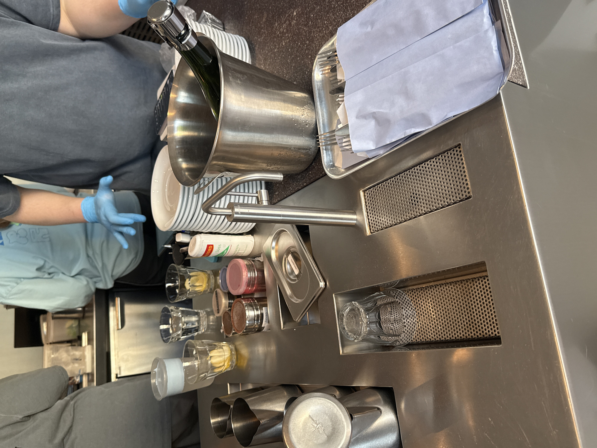 The image displays an indoor scene, likely a service counter, bar, or professional kitchen, characterized by extensive use of stainless steel equipment. Two individuals are partially visible, both wearing light blue disposable gloves, indicating a focus on hygiene, common in food and beverage preparation or service.
In the central foreground, a large, cylindrical stainless steel ice bucket contains a green glass bottle, likely champagne. To its left, a stack of white plates is neatly arranged. A modern, industrial-style chrome-plated faucet with an articulated arm is prominent on the counter. Adjacent to the ice bucket, a rectangular metal tray holds various pieces of cutlery, individually wrapped in light blue paper napkins.
The stainless steel countertop integrates several functional elements. There are recessed areas holding clear glasses, some containing yellow lemon slices. Alongside these are small, transparent containers with different colored powdered contents, possibly spices or cocktail ingredients. A white bottle, partially visible, bears the text "FORD" and "Biss" on its label. Another small, lidded metal container, possibly for ingredients, has "Mozzarella" faintly inscribed on its lid. Further down the counter, several stainless steel pitchers or frothing jugs are neatly placed in dedicated slots. Perforated metal grates are integrated into the counter surface, likely for drainage or air circulation.
One person, wearing a dark grey t-shirt and blue gloves, is positioned near the ice bucket. Another person, dressed in a light blue top and blue gloves, is visible in the background. The setup suggests an environment dedicated to efficient beverage and possibly food preparation or serving. No specific visual cues within the image indicate the location as Moscow, Russia. The time of day and the individuals' emotions are not discernible.