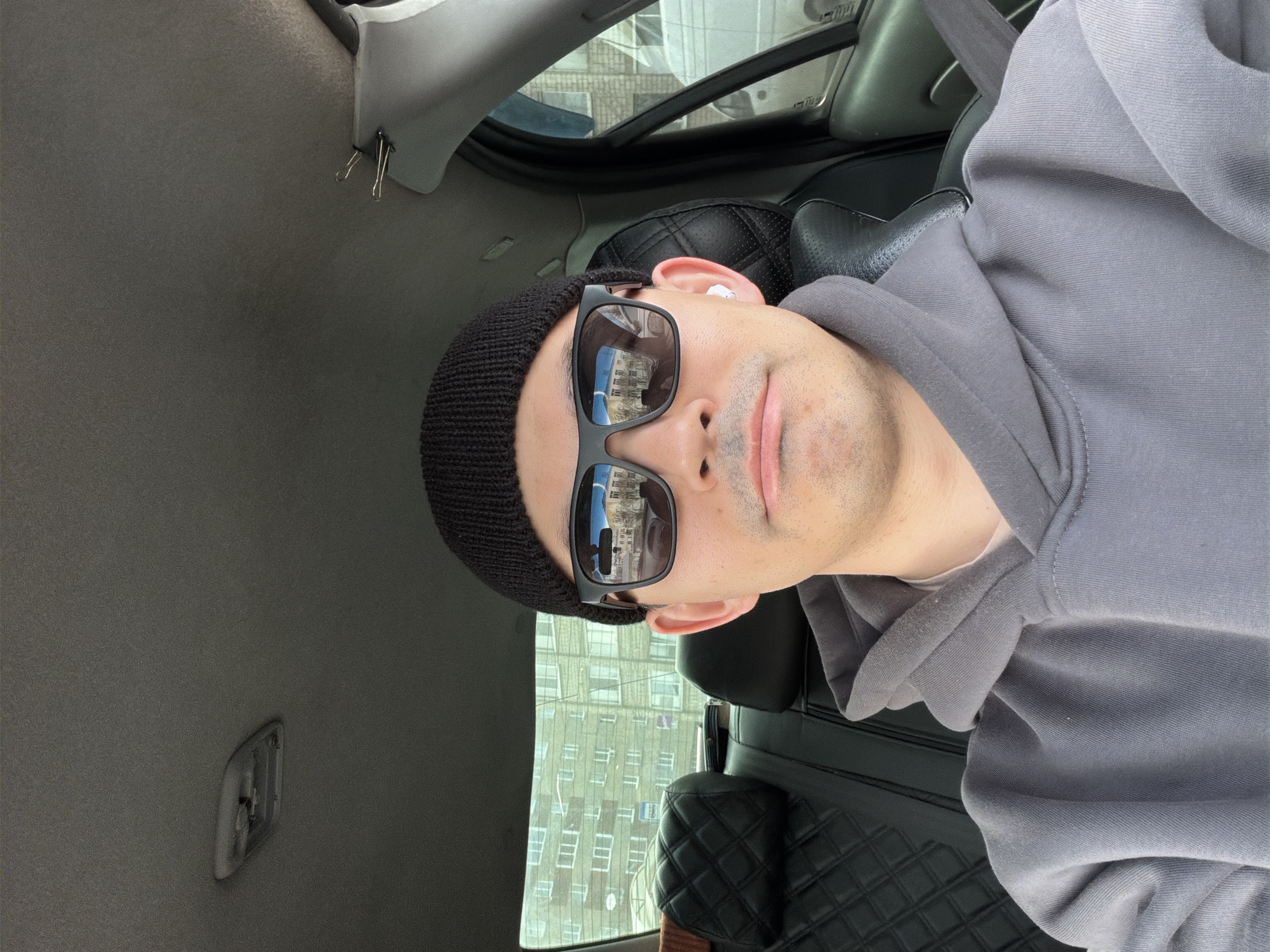 A man wearing a black beanie and sunglasses sits in a car. The reflection in his sunglasses shows buildings, indicating an urban environment. He is wearing a grey hoodie. The car's interior is visible, including the headliner, seat, and window. The reflections in the glasses appear to show a location in Izhevsk, Russia, based on the architectural style of the buildings in the reflection.