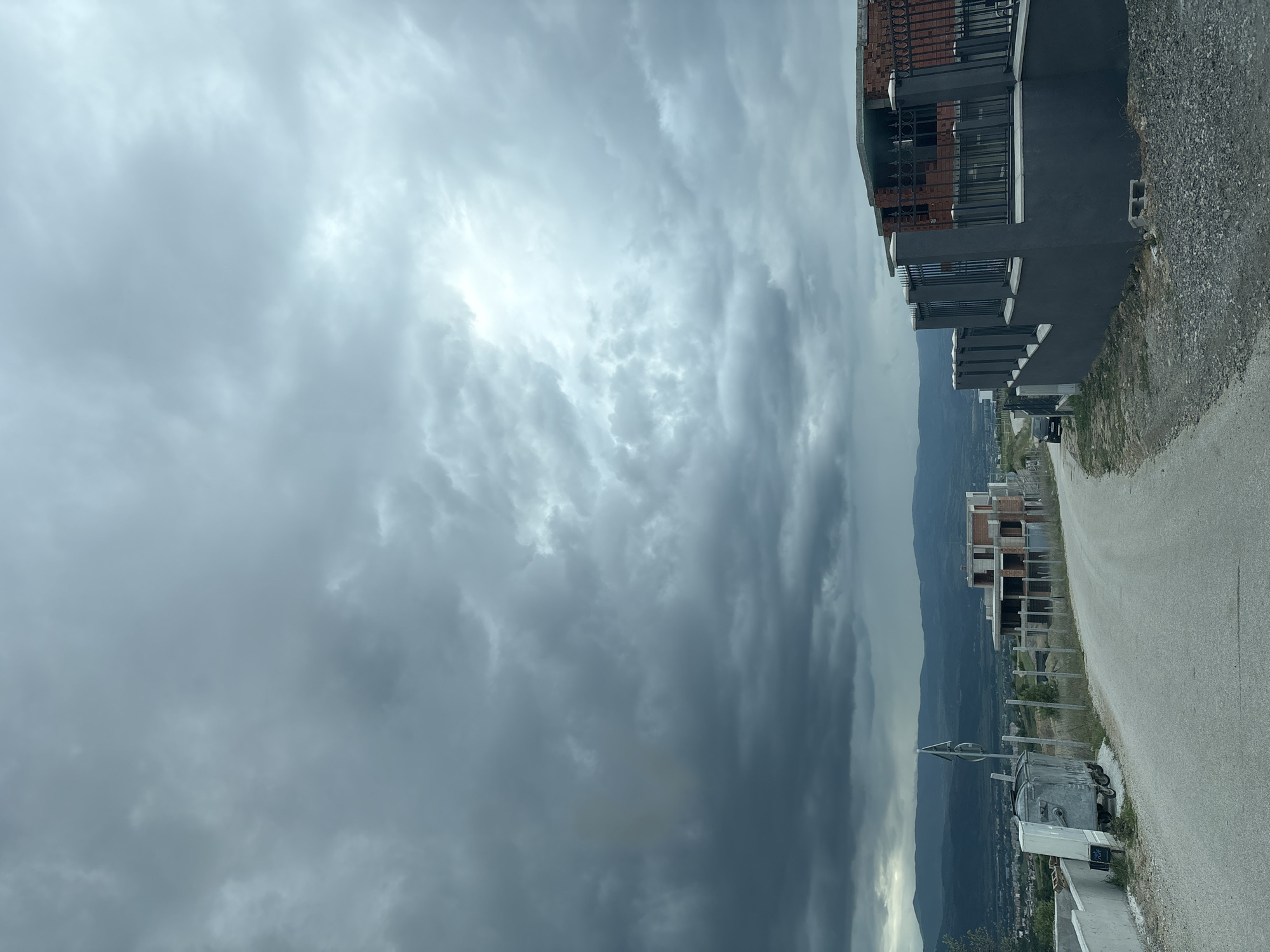 The image shows a cloudy sky above a road with buildings on either side. The road is paved and appears to be sloping downwards. The buildings on the right side of the road are modern and appear to be under construction or recently built. In the distance, there are mountains and what looks like a valley or a plain. A large trash container is along the road. The sky is overcast with dark, dramatic clouds. The location is in an unidentified area within the city of Unknown, Unknown.