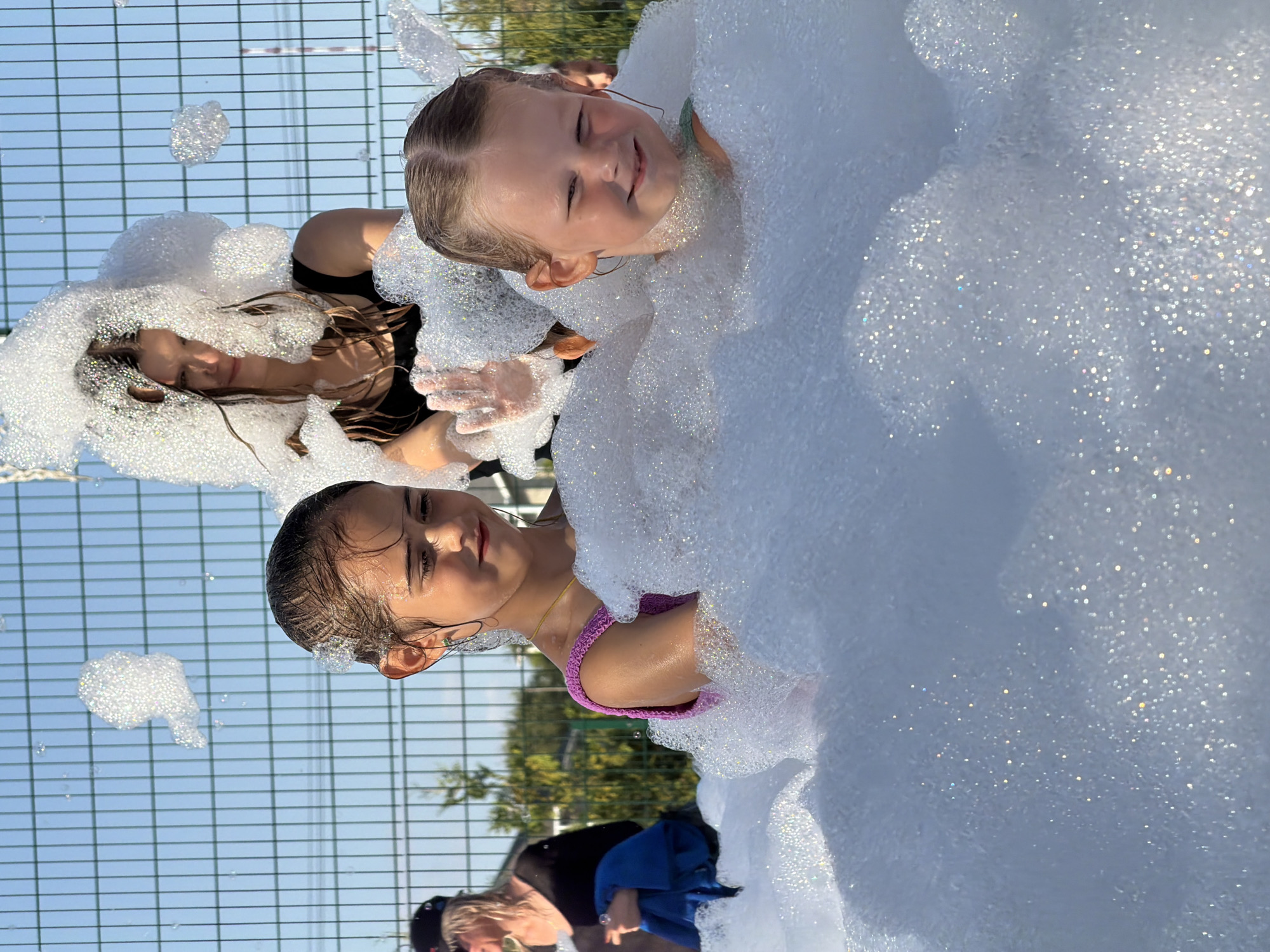 The image depicts a sunny outdoor scene, likely an event involving a foam machine. Two young girls are prominently featured, submerged in a large amount of white foam. The girl on the right, with blonde hair pulled back, is smiling and looking towards the viewer, wearing a green top. The girl on the left, with dark hair, is looking to her right, also smiling and appears to be wearing a pink top. Behind them, a young woman with dark hair, wearing a black top, is visible, her face partially obscured by foam. She is holding up a hand, fingers spread. In the background, a green fence runs horizontally, with trees and sky visible beyond. A woman wearing a black shirt and a black hat is visible to the far left, holding a blue item. The overall impression is one of fun and enjoyment in a foamy environment, possibly a park or recreational area within the city of Kamyshin, Russia.