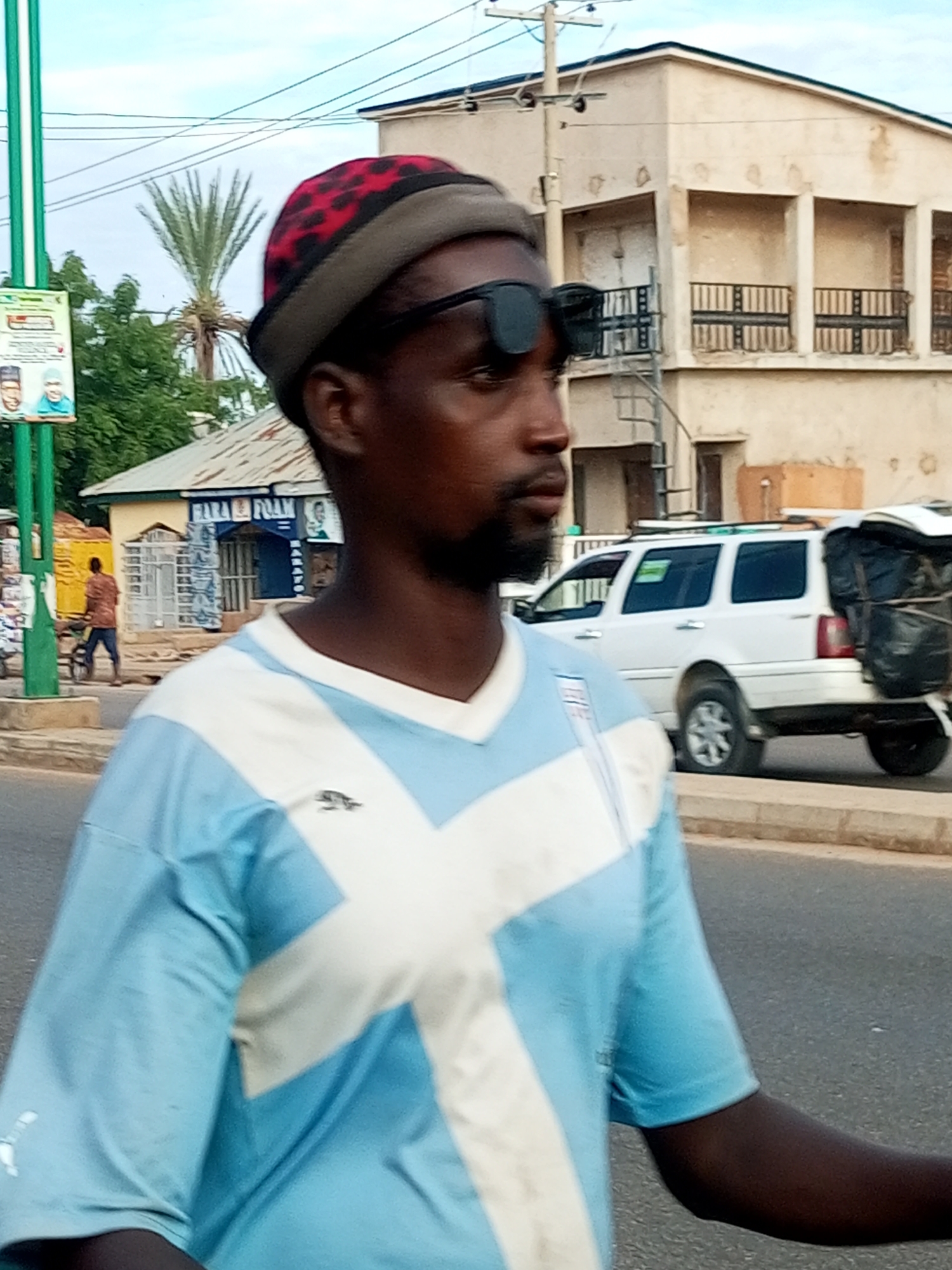 A dark-skinned man occupies the foreground, facing right with his head slightly turned, suggesting a profile view. He wears a light blue short-sleeved shirt with a prominent white 'X' pattern on the front, a small dark animal logo on the left chest, and a white 'Puma' logo on the left sleeve. His head is covered by a dark grey beanie with red and black leopard print accents. A black eye patch is worn over his right eye, with dark sunglasses resting on top of the eye patch. He has a short beard or stubble.
The background reveals a bustling street scene. To the left, a tall green pole with a white and green sign is visible; the sign displays two male faces, possibly political figures, at the bottom. Behind the pole, a distinct palm tree stands against a light blue sky with sparse clouds. Further back, a low-rise building with a corrugated metal roof, painted light blue and yellow, has a sign that appears to include "HARA FOAM". A man in a brown shirt and blue pants walks away from the camera, pushing a bicycle or cart on the paved road.
To the right of the foreground subject, a multi-story building with a worn, light-colored facade features multiple recessed balconies with black metal railings. Utility poles and wires are visible against the sky. On the paved road, a white SUV is seen from the rear-right, with black items, possibly luggage or goods, secured to its back. The road is asphalt, bordered by a grey concrete curb.