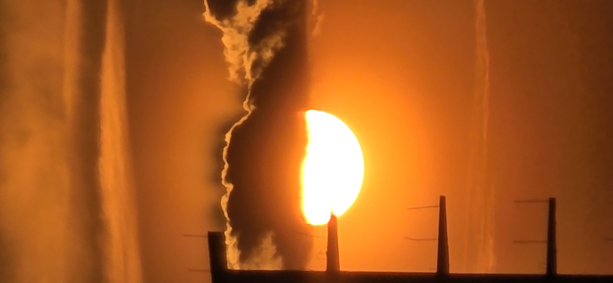 A close-up captures a striking sunset in Bar, Montenegro. The sun, appearing as a partially obscured bright orb, radiates an intense orange glow that permeates the scene. A dark, silhouetted structure with posts is in the foreground. The sky is filled with an orange fog or haze. To the left of the sun, a cloud or smoke formation partially obscures the light.
